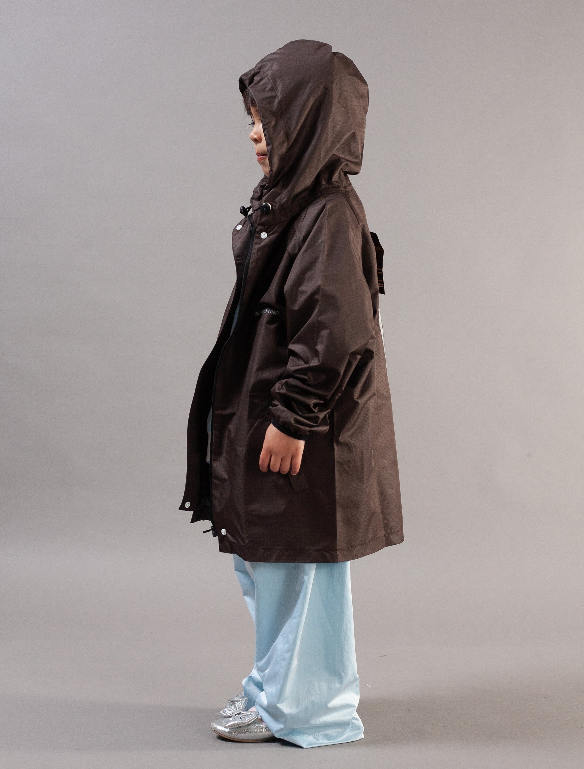 A child wearing a dark brown hooded raincoat, light blue pants, and silver sneakers, shown in profile against a plain gray studio background.