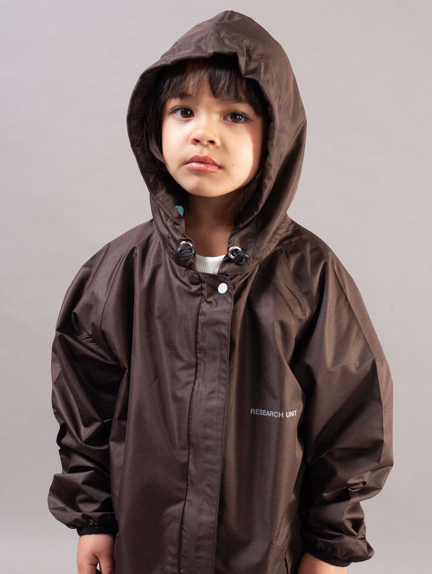 A child wearing a dark brown hooded rain jacket with a white logo on the chest, photographed against a plain gray studio background.