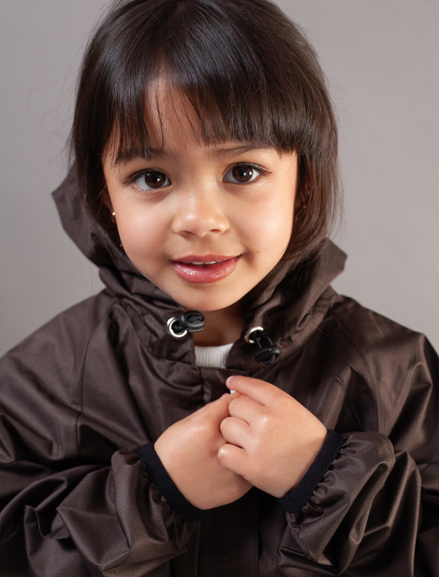 A young child wearing a dark brown hooded rain jacket, smiling at the camera against a plain gray studio background.