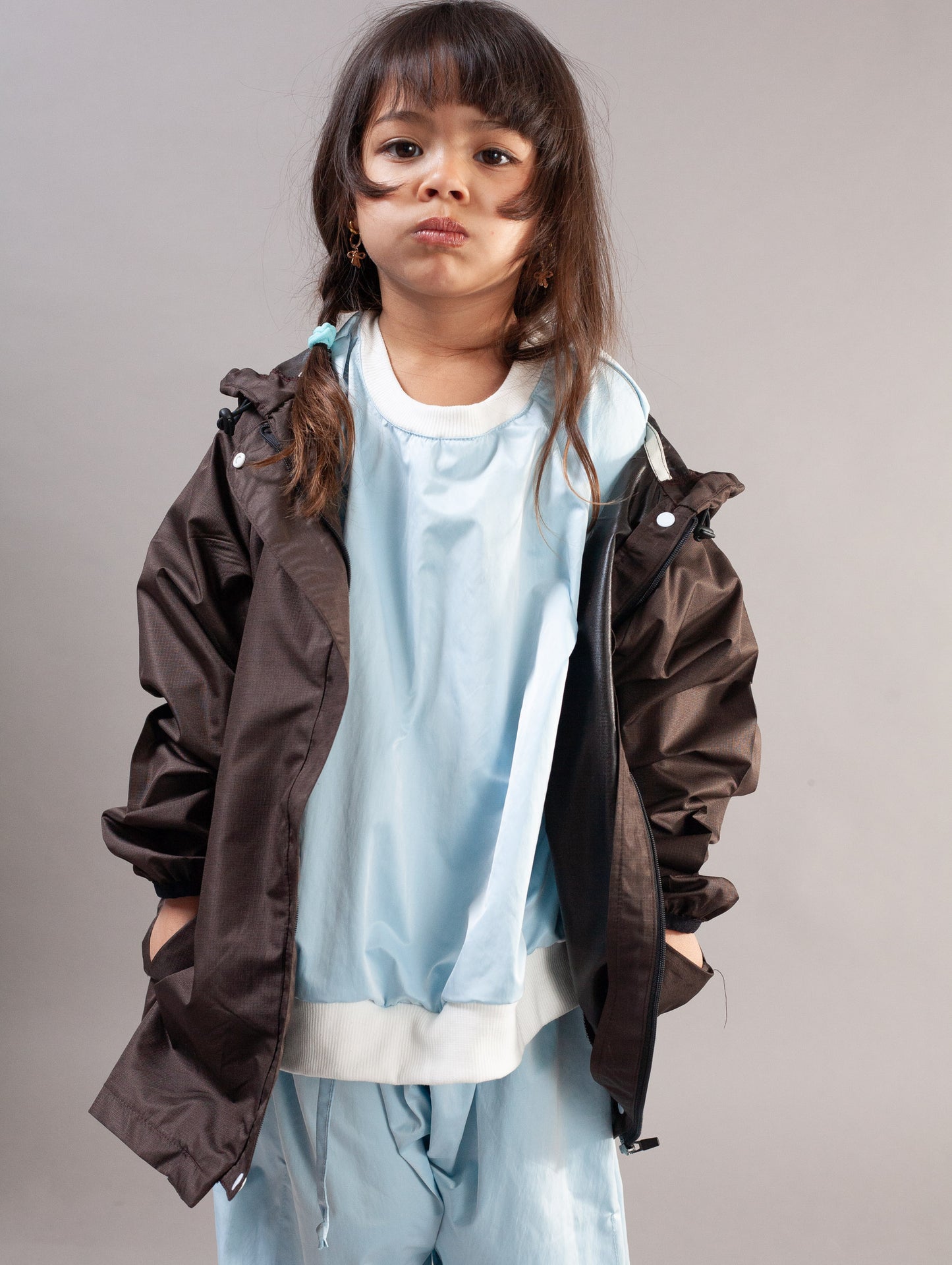 A young girl modeling a dark brown lightweight jacket over a light blue sweatshirt and matching light blue pants, photographed against a solid gray studio background.