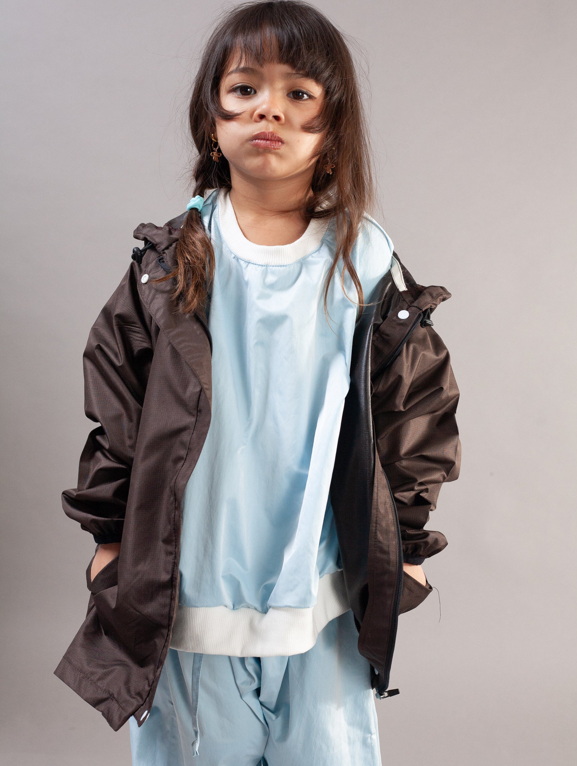 A young girl modeling a dark brown lightweight jacket over a light blue sweatshirt and matching light blue pants, photographed against a solid gray studio background.