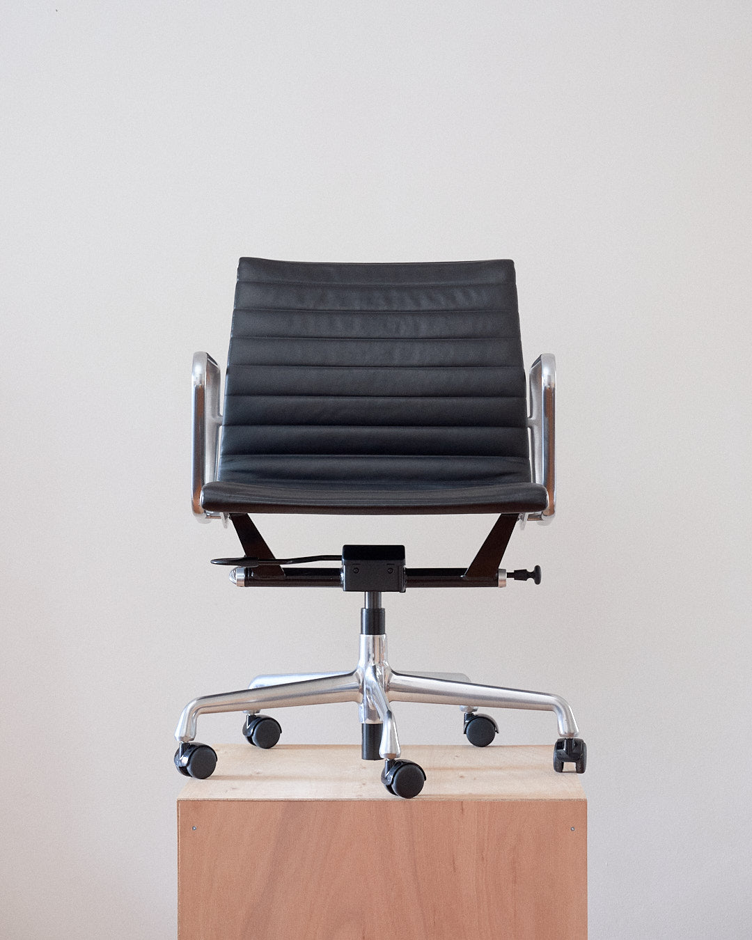 A modern black leather office chair with a ribbed backrest, chrome metal armrests, and a five-star wheeled base, shown sitting on a light wood pedestal against a plain off-white wall.