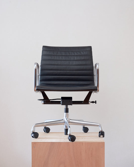 A modern black leather office chair with a ribbed backrest, chrome metal armrests, and a five-star wheeled base, shown sitting on a light wood pedestal against a plain off-white wall.