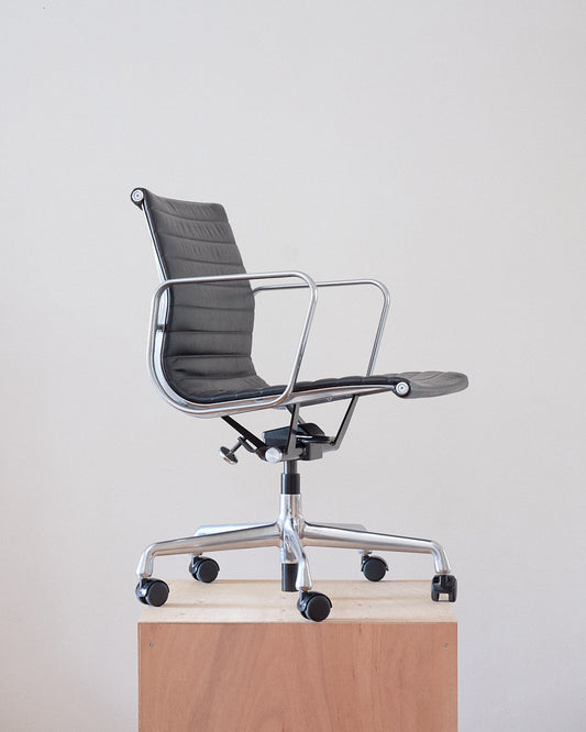 A modern black leather office chair with a chrome metal frame and five-wheel base, shown on a light wood pedestal against a plain off-white background.