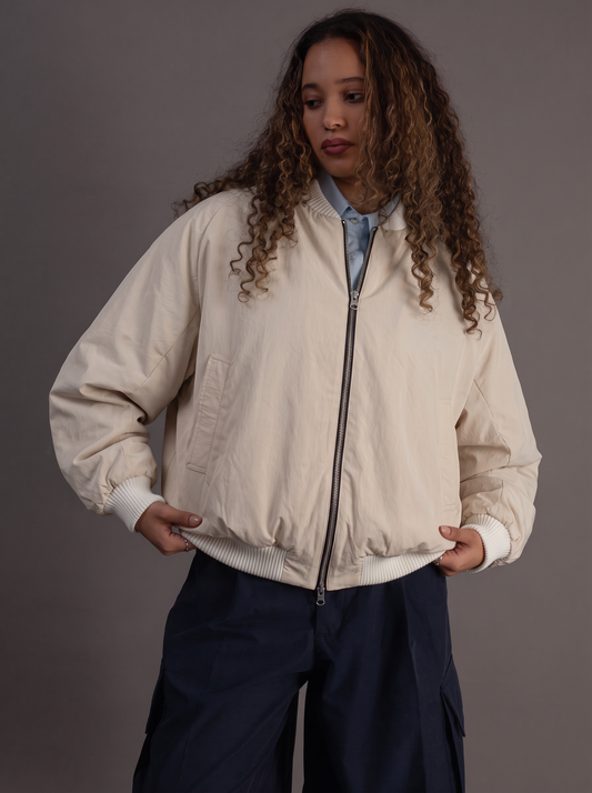 A woman with long curly brown hair wearing a cream-colored zip-up bomber jacket over a light blue collared shirt and dark navy cargo pants, posed against a solid dark gray studio background.