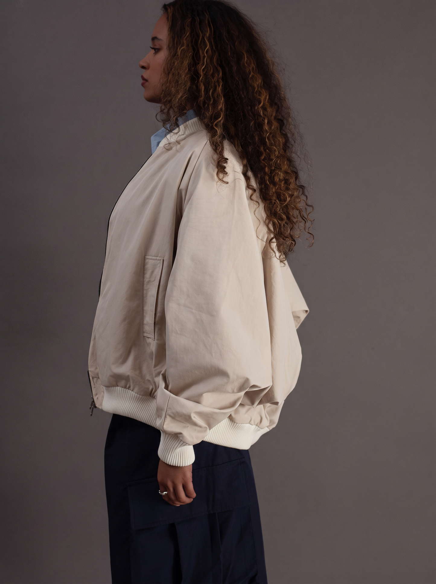 A woman shown in side profile wearing a cream-colored oversized bomber jacket with voluminous sleeves and dark navy pants, set against a solid dark gray studio background.