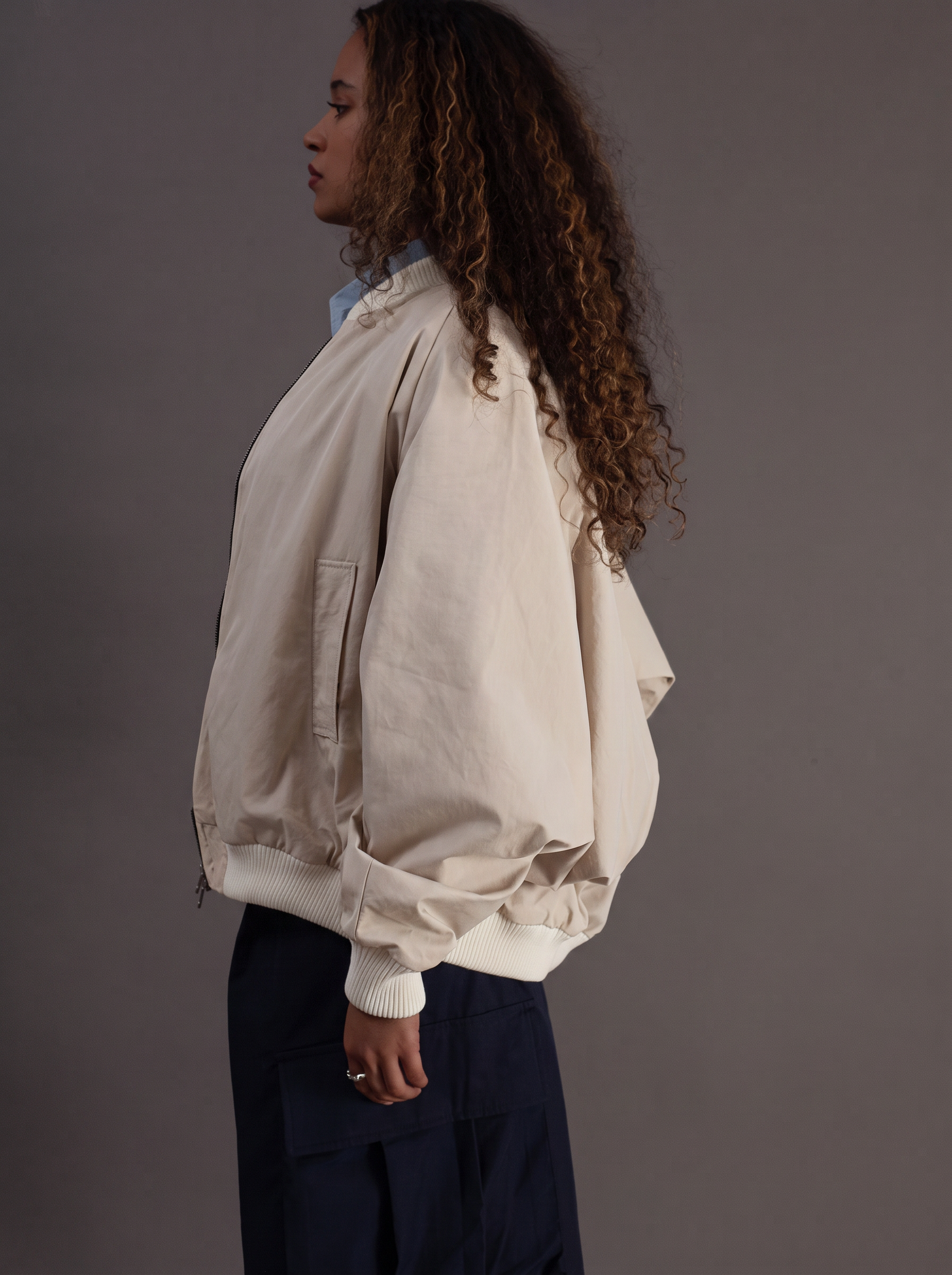 A woman shown in side profile wearing a cream-colored oversized bomber jacket with voluminous sleeves and dark navy pants, set against a solid dark gray studio background.
