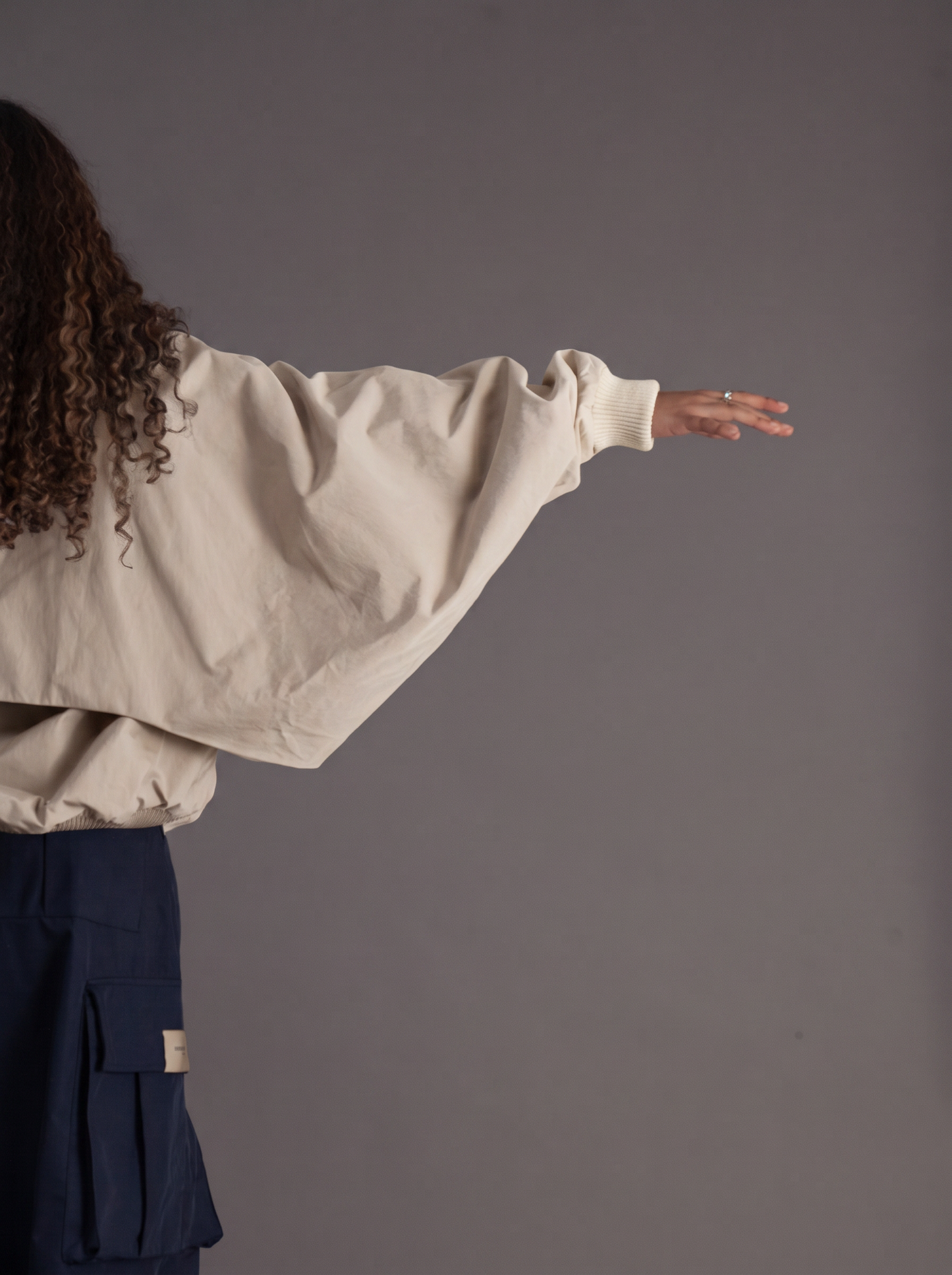 A person shown from the back wearing a cream-colored oversized long-sleeve top and dark navy cargo pants, extending their right arm against a solid dark gray studio background.