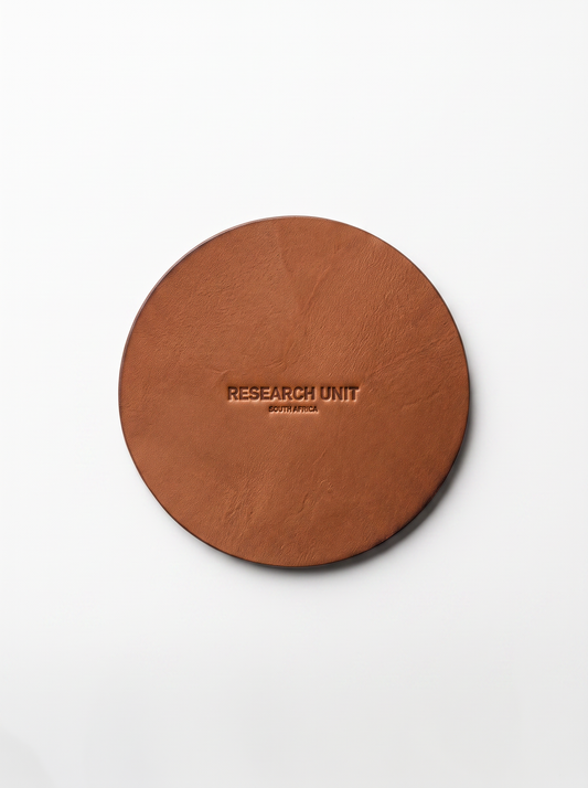 A round brown leather coaster with 'RESEARCH UNIT' embossed in the center, photographed from directly above on a clean white background.
