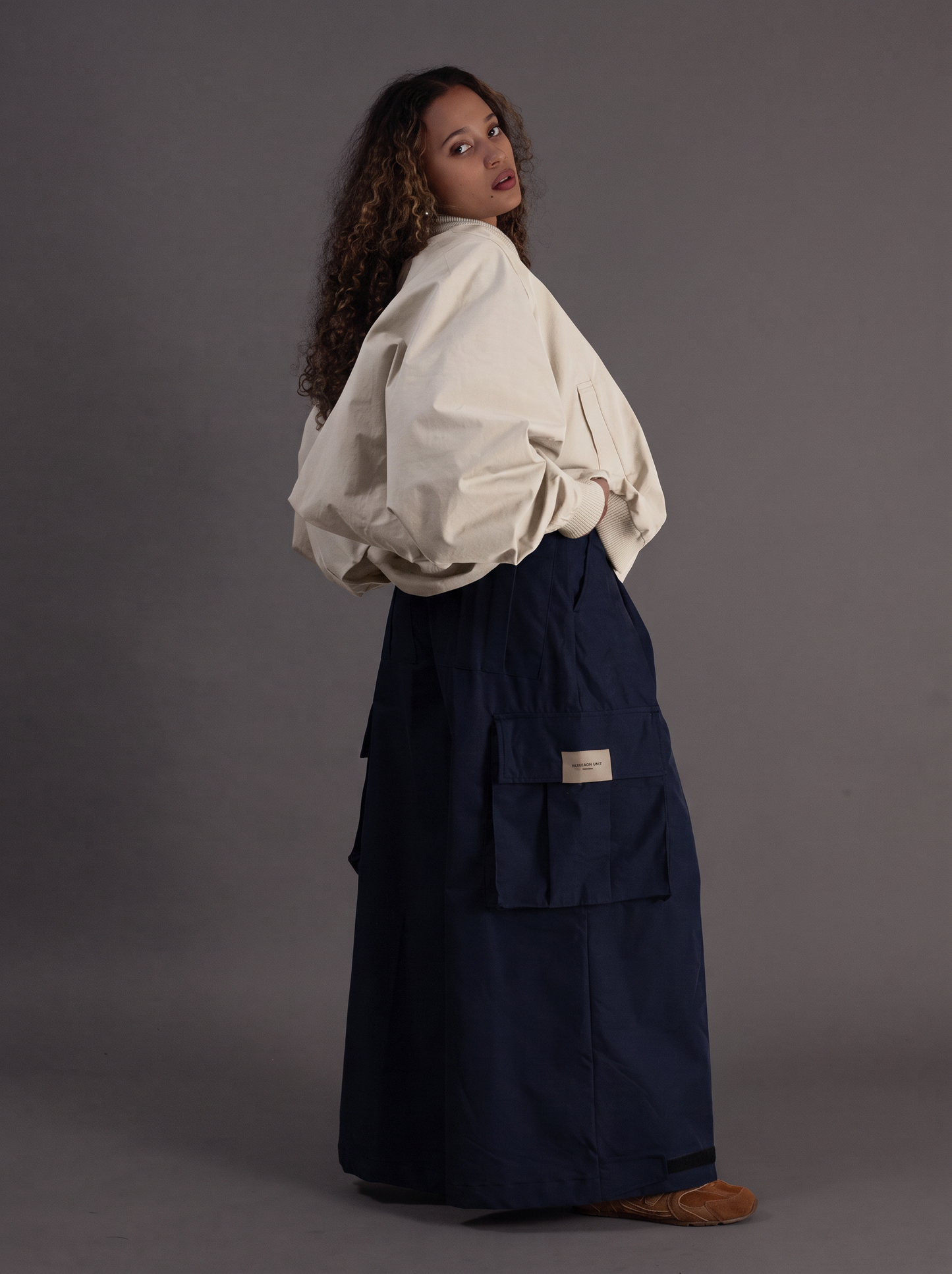 A model wearing a cream-colored oversized long-sleeve blouse and a dark navy blue cargo skirt, standing against a solid gray studio background.