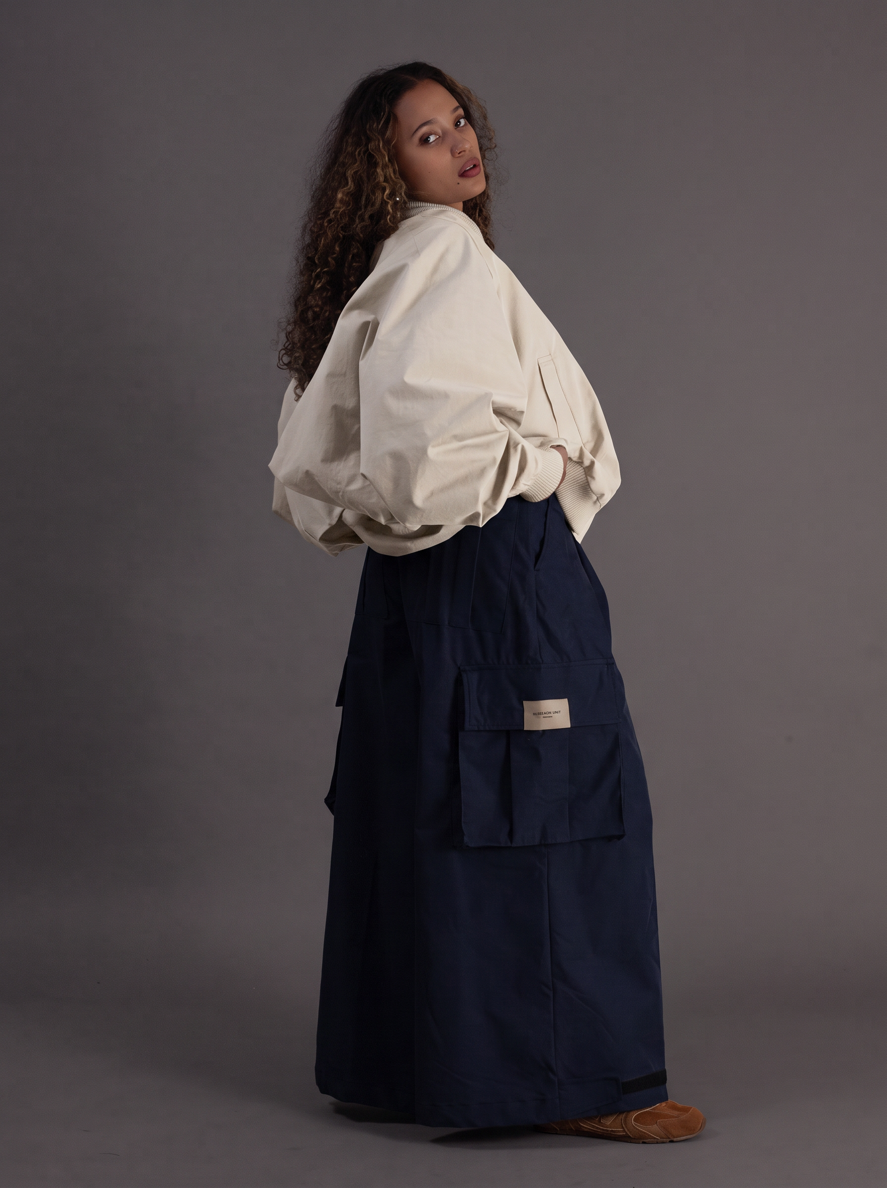 A model wearing a cream-colored oversized long-sleeve blouse and a dark navy blue cargo skirt, standing against a solid gray studio background.