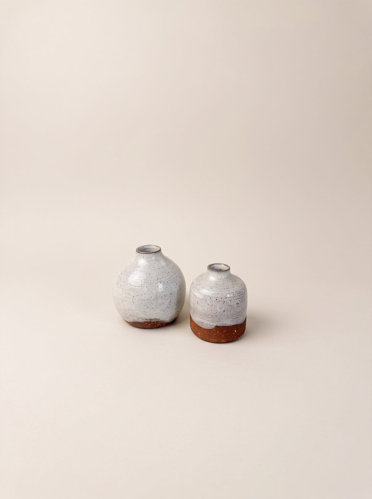 Two small ceramic vases with rounded bulbous bodies, speckled light gray glazes, and exposed terracotta bases, photographed on a plain warm beige studio background.