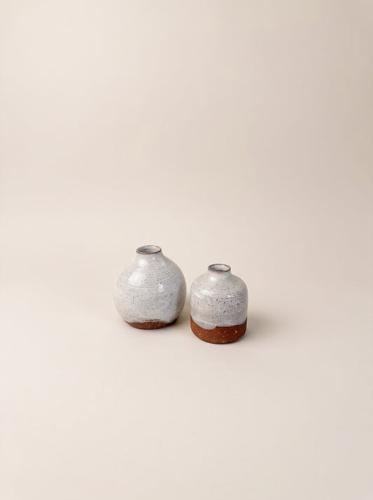 Two small ceramic vases with rounded bulbous bodies, speckled light gray glazes, and exposed terracotta bases, photographed on a plain warm beige studio background.