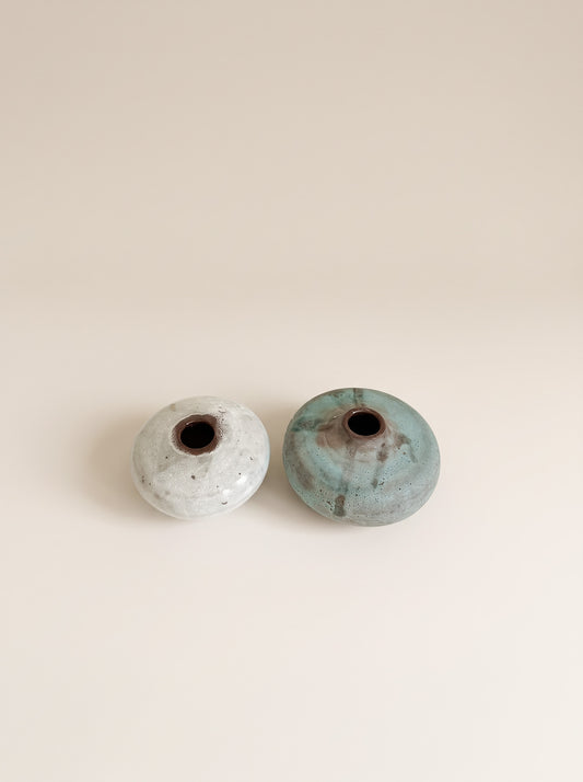 Two small round ceramic vases with open centers, one in a light speckled off-white glaze and the other in a mottled teal-green glaze, set against a solid warm beige background.