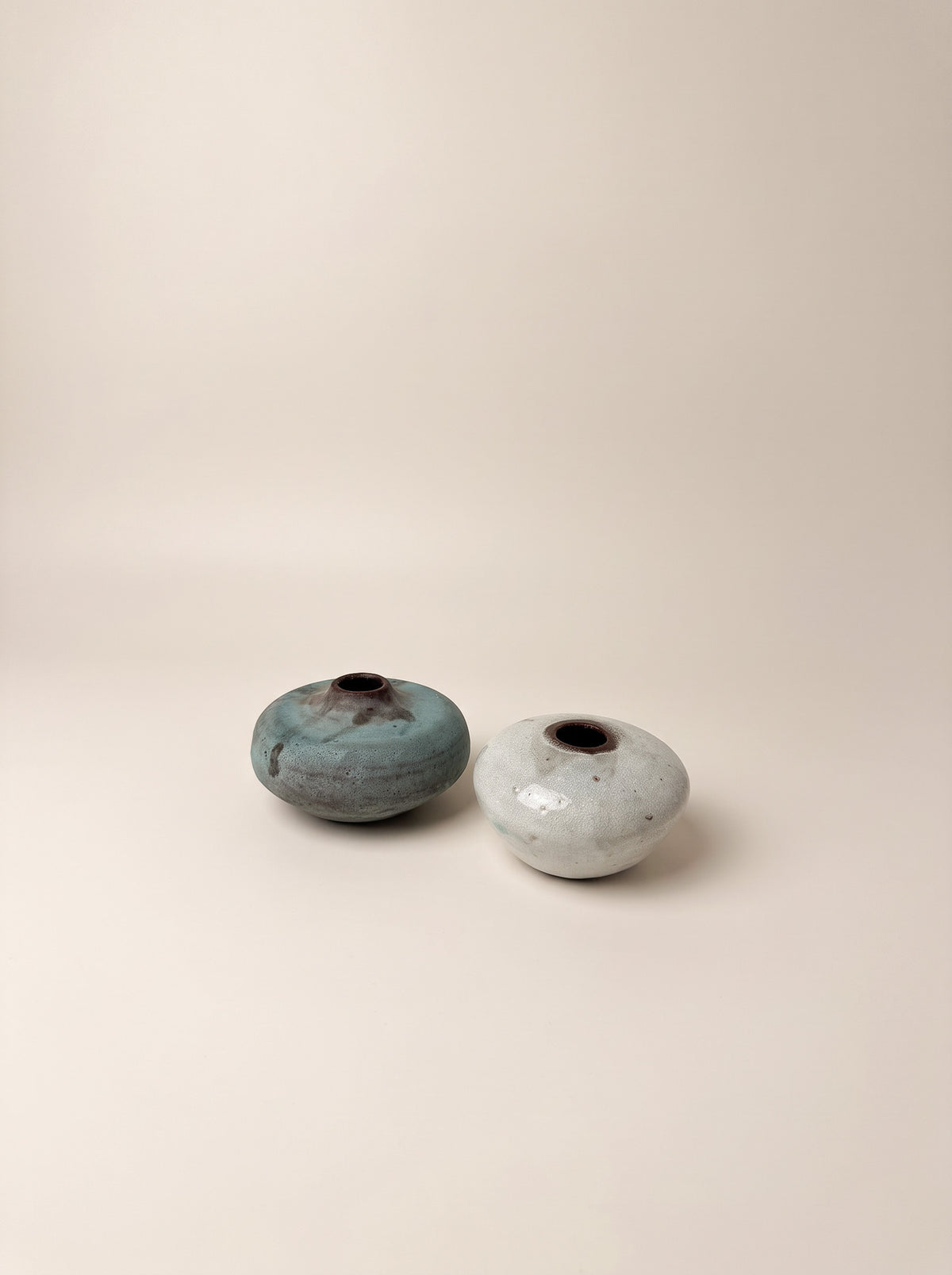 Two small handmade ceramic vases with rounded, egg-shaped bodies and small dark openings, displayed on a warm beige studio background. One vase has a textured teal-blue glaze with brown speckling, and the other has a speckled off-white glaze.