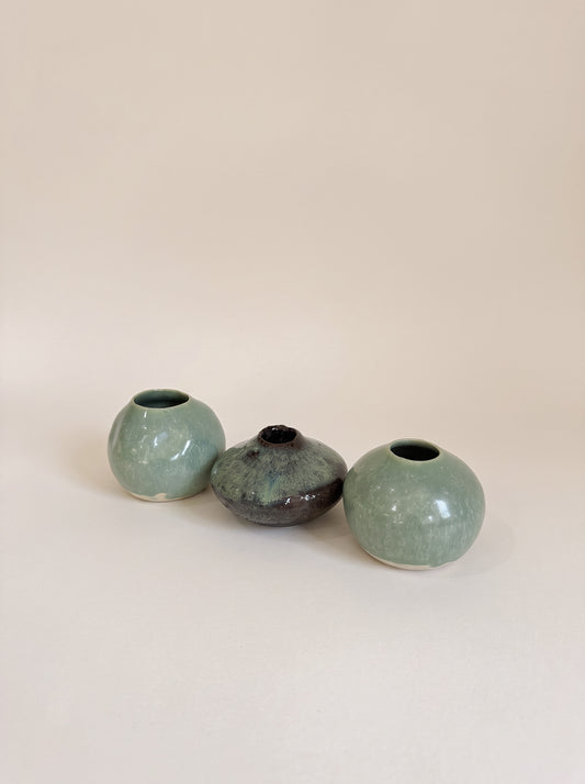 Three small ceramic vases in a row on a plain beige background. The left and right vases are a pale mint green glaze, and the center vase is a dark, mottled charcoal gray with subtle blue-green undertones.