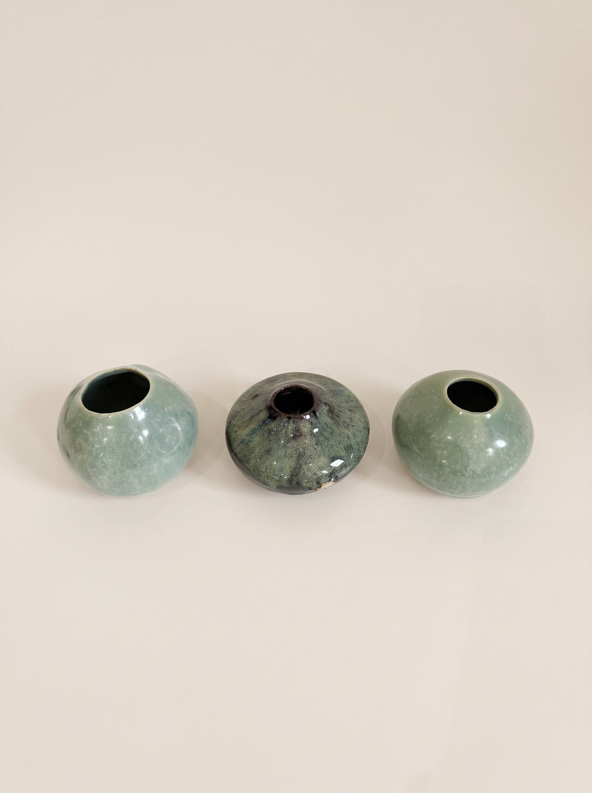 Three small round ceramic vases in a row on a plain light beige background. The left and right vases are a pale mint green with a glossy finish, while the center vase has a mottled dark green and black glaze.