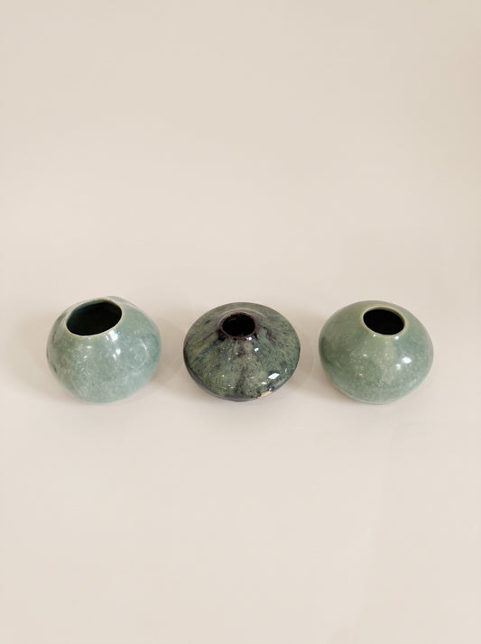 Three small round ceramic vases in a row on a plain light beige background. The left and right vases are a pale mint green with a glossy finish, while the center vase has a mottled dark green and black glaze.