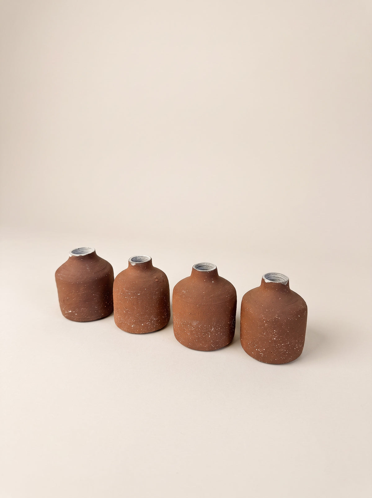Four small terracotta-colored ceramic bottles with silver caps lined up in a row on a warm beige background.