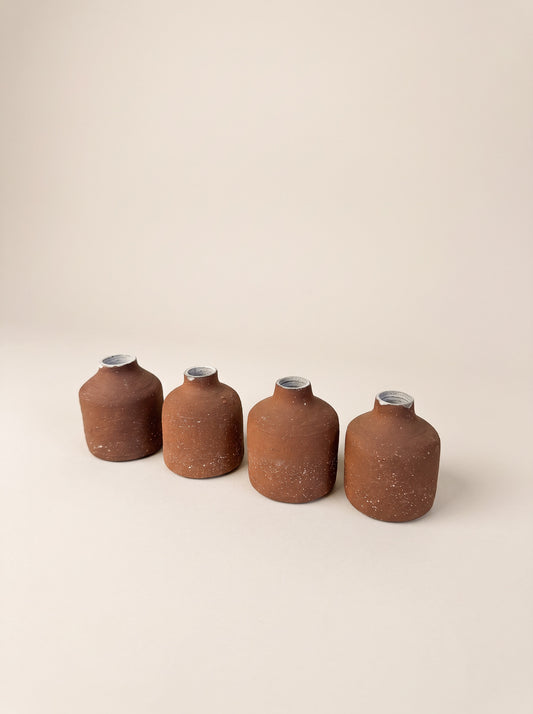 Four small terracotta-colored ceramic bottles with silver caps lined up in a row on a warm beige background.