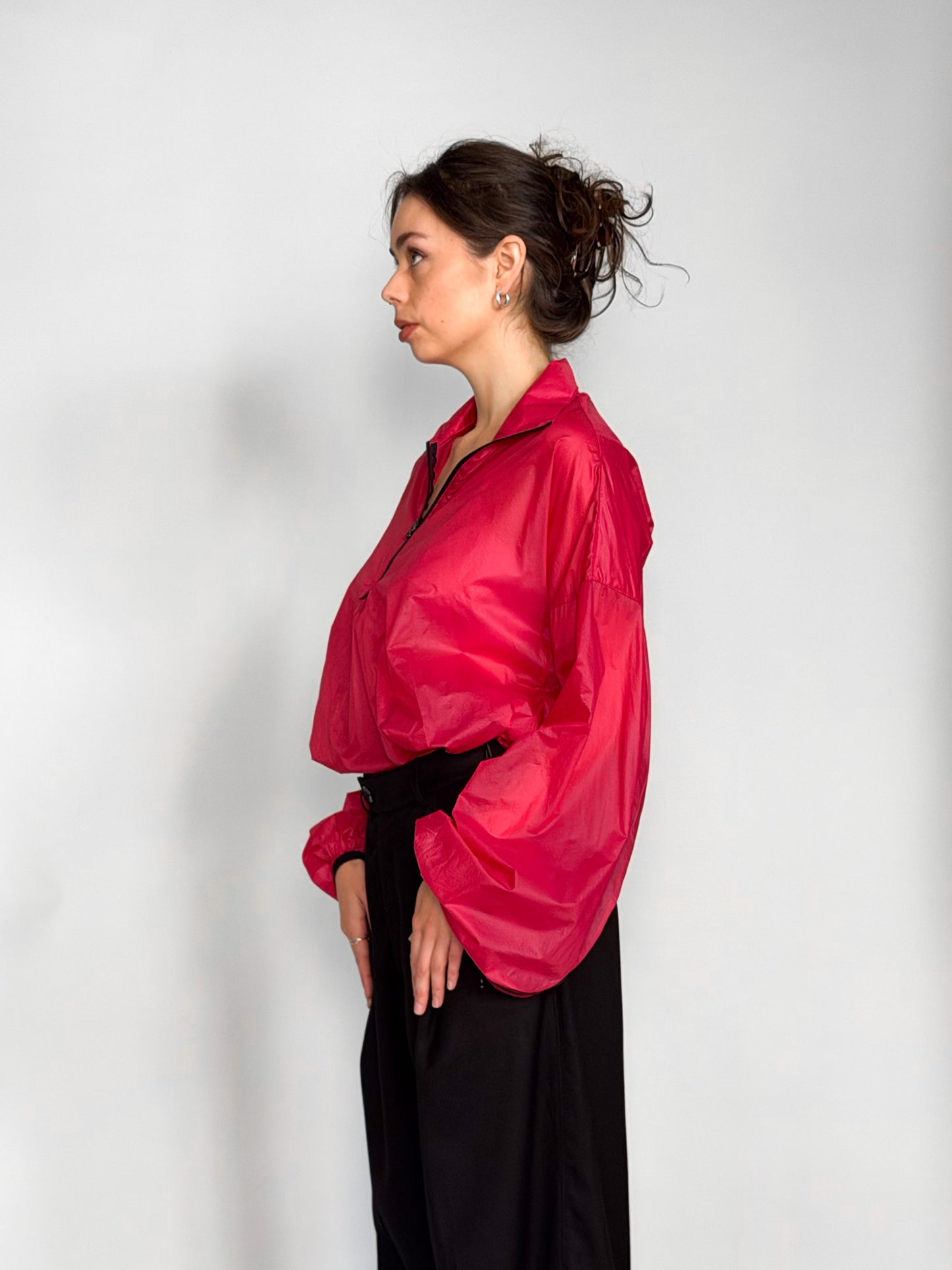 A woman modeling a bright pink semi-sheer zip-up windbreaker jacket with voluminous balloon sleeves, paired with black wide-leg trousers, against a light gray studio background.
