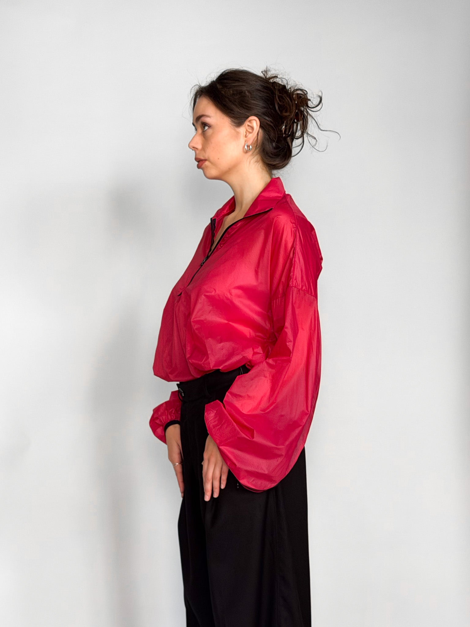 A woman modeling a bright pink semi-sheer zip-up windbreaker jacket with voluminous balloon sleeves, paired with black wide-leg trousers, against a light gray studio background.