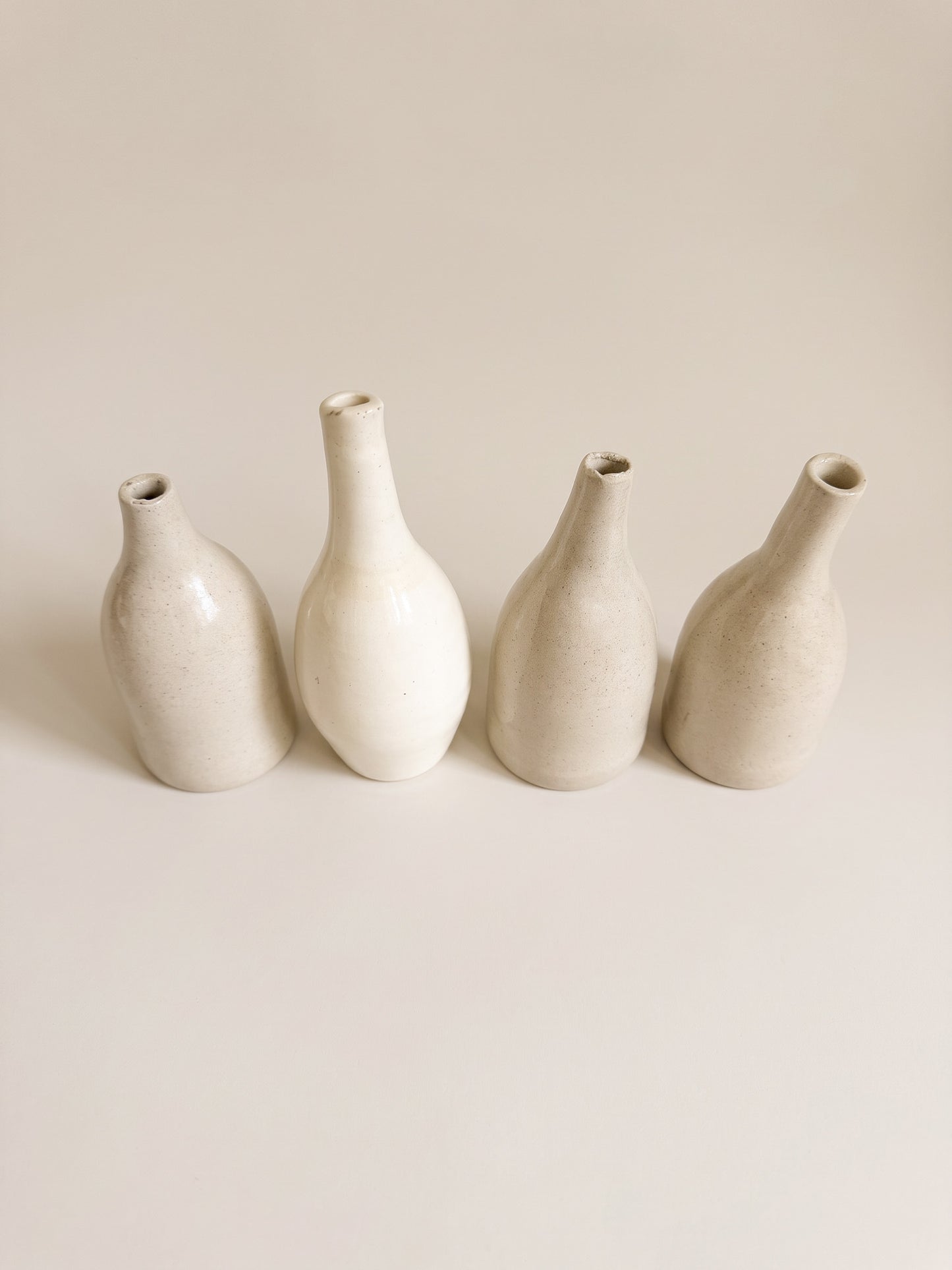 Four small ceramic vases in a minimalist studio setting, arranged in a row against a warm off-white background.