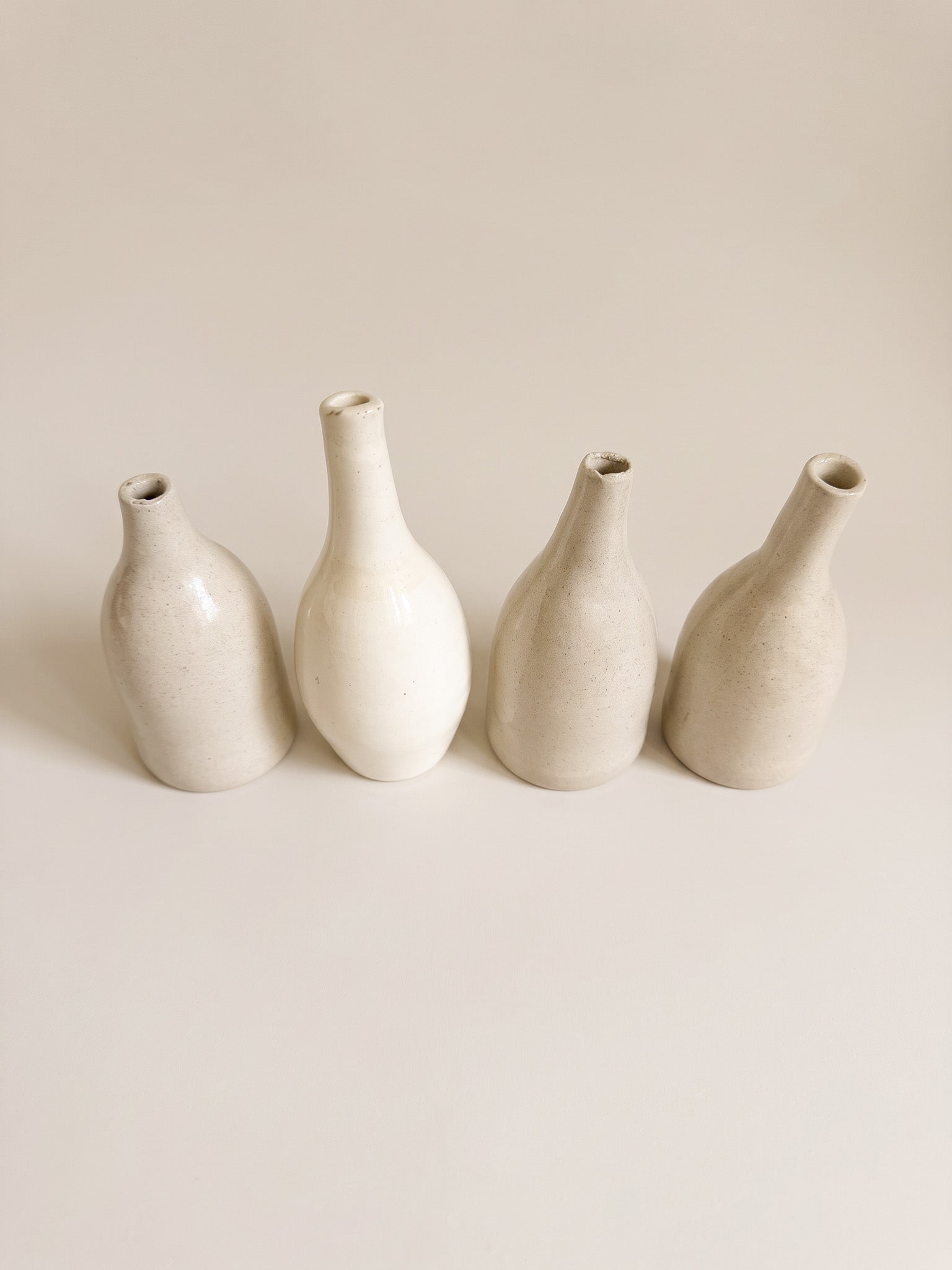 Four small ceramic vases in a minimalist studio setting, arranged in a row against a warm off-white background.