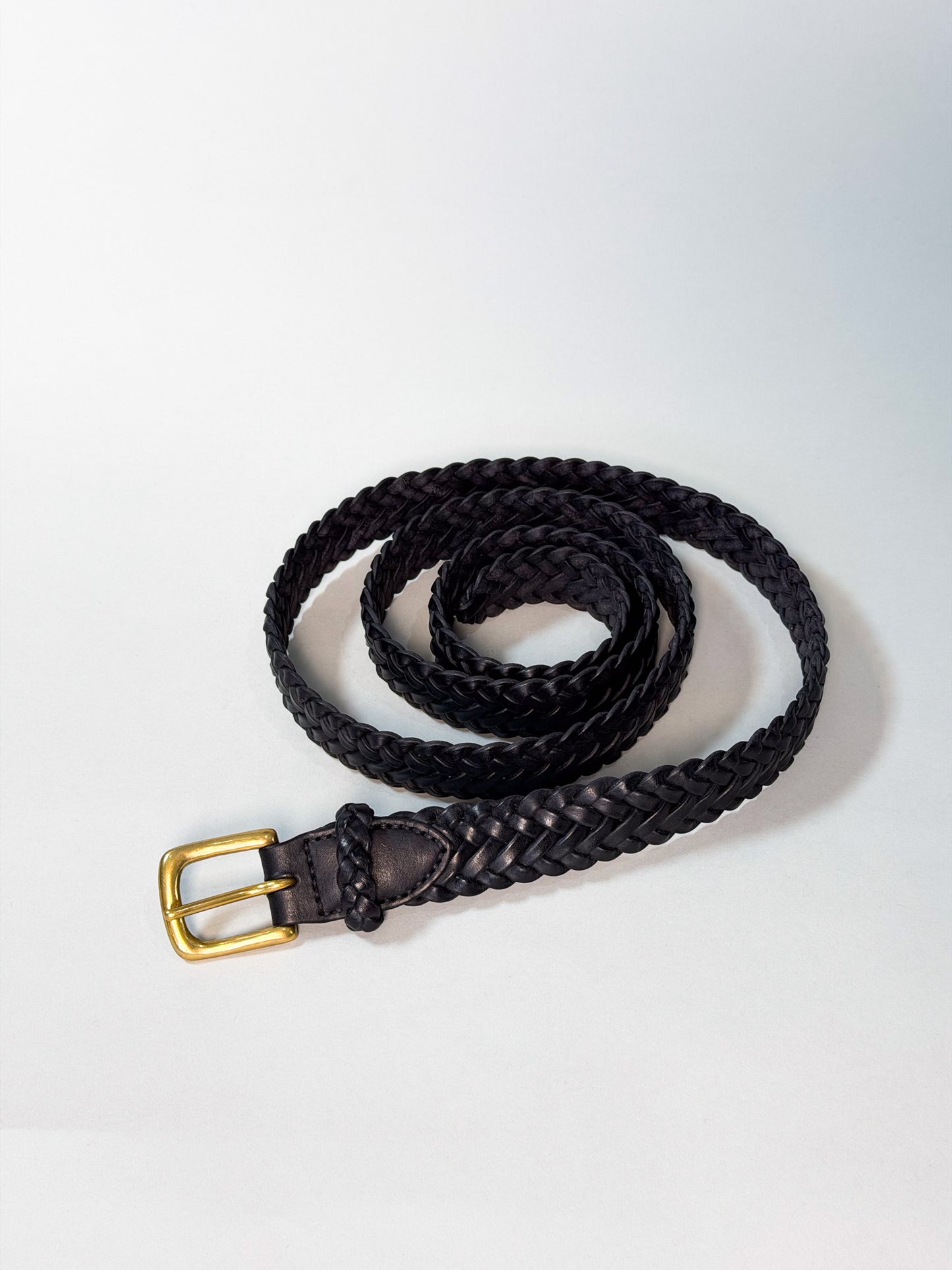 A black braided leather belt with a gold-tone metal buckle, coiled on a light gray background.