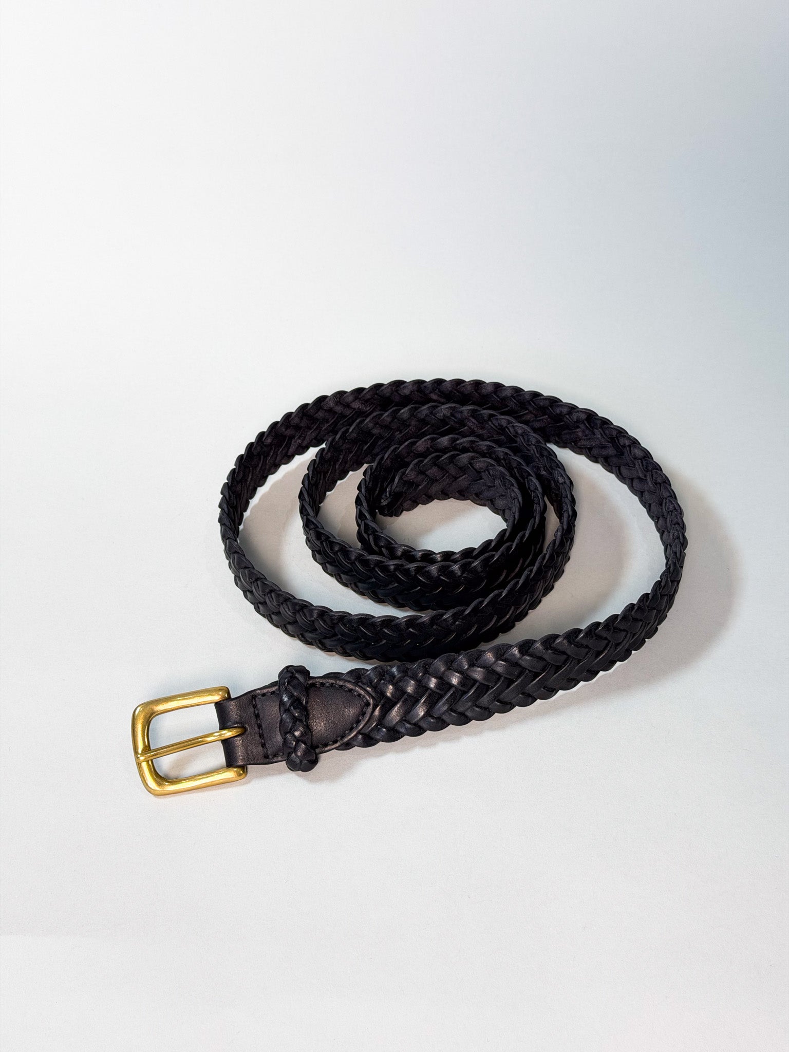 A black braided leather belt with a gold-tone metal buckle, coiled on a light gray background.