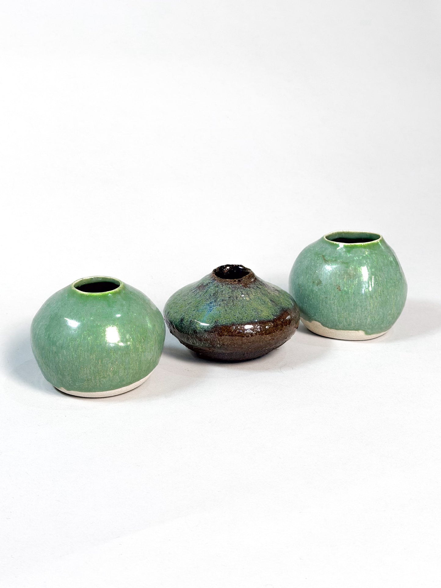 Three handmade ceramic vases with a glossy glaze, displayed on a clean white background. Two vases are a light green color with a white base, and the center vase is a dark brown with a green and blue iridescent glaze.