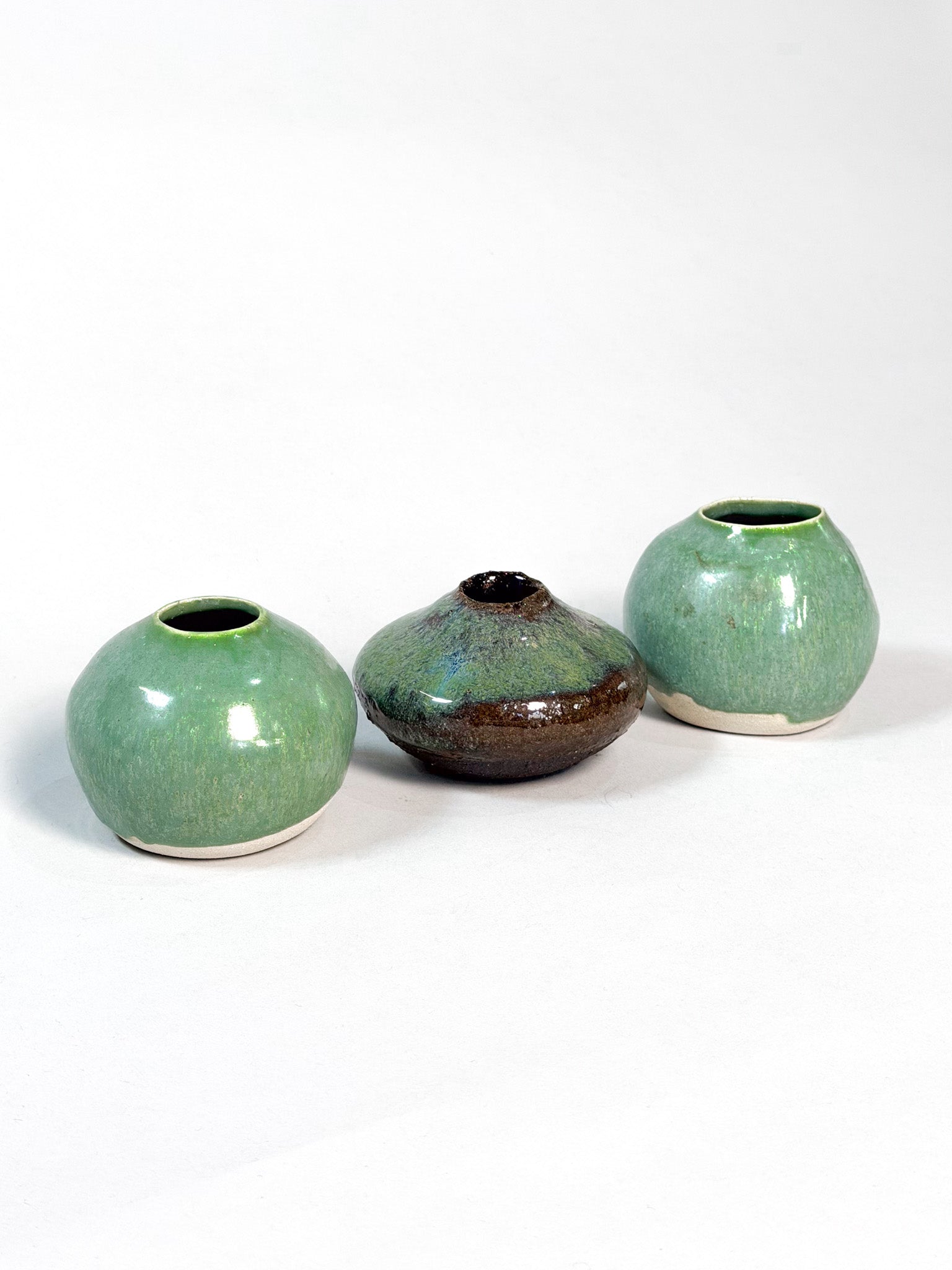 Three handmade ceramic vases with a glossy glaze, displayed on a clean white background. Two vases are a light green color with a white base, and the center vase is a dark brown with a green and blue iridescent glaze.