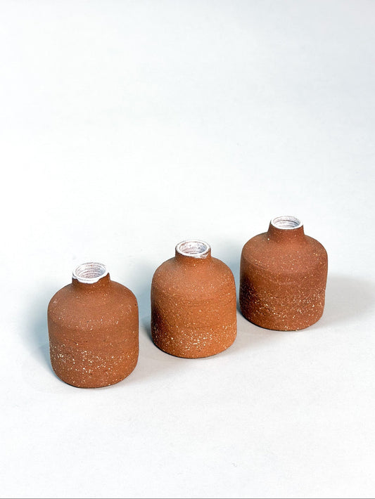 Three small terracotta-colored ceramic bottles with textured surfaces and silver metal collars, arranged in a row on a light gray background.