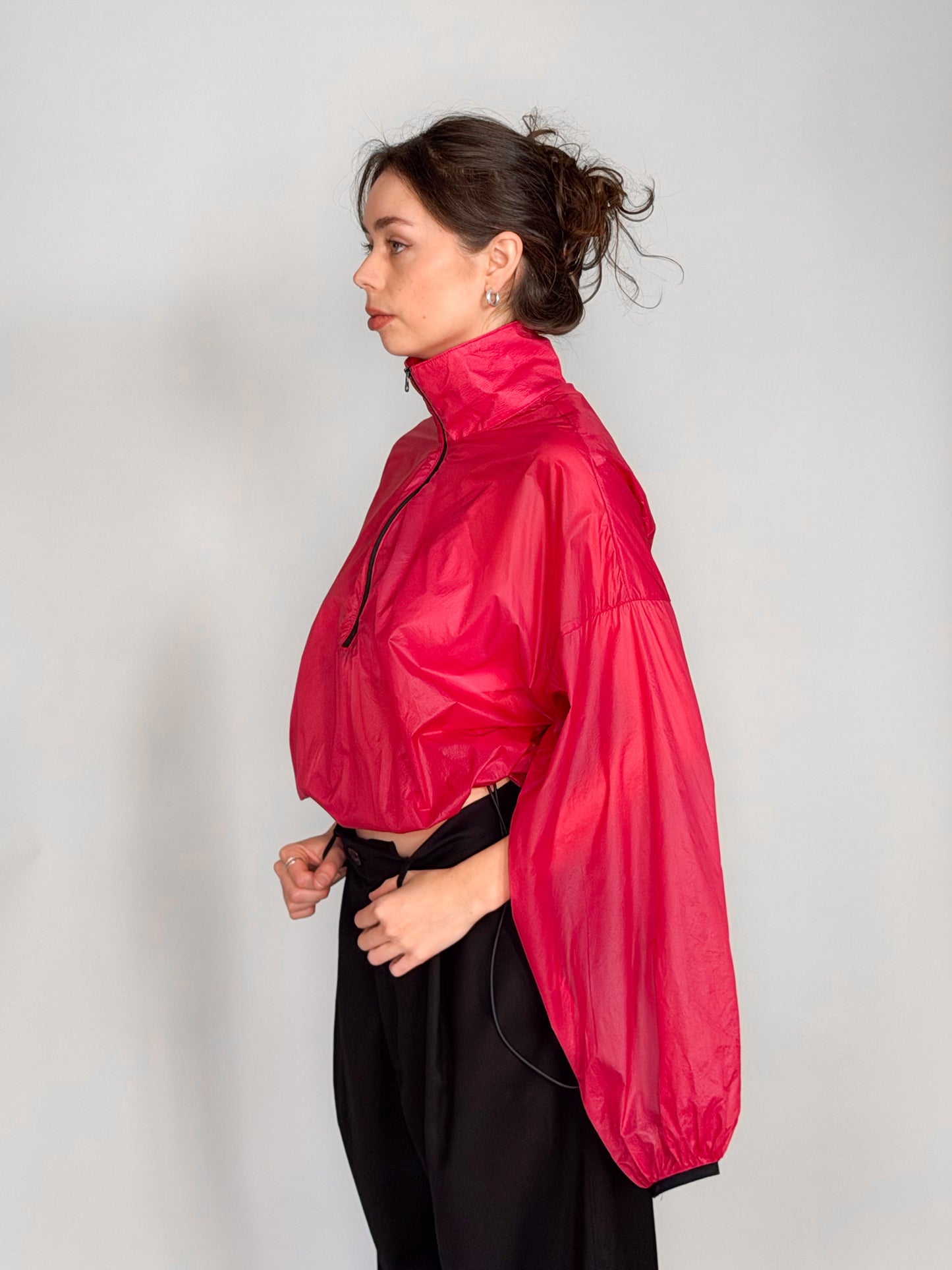 A woman modeling a bright pink semi-sheer zip-up windbreaker jacket with voluminous balloon sleeves, paired with black wide-leg trousers, against a light gray studio background.