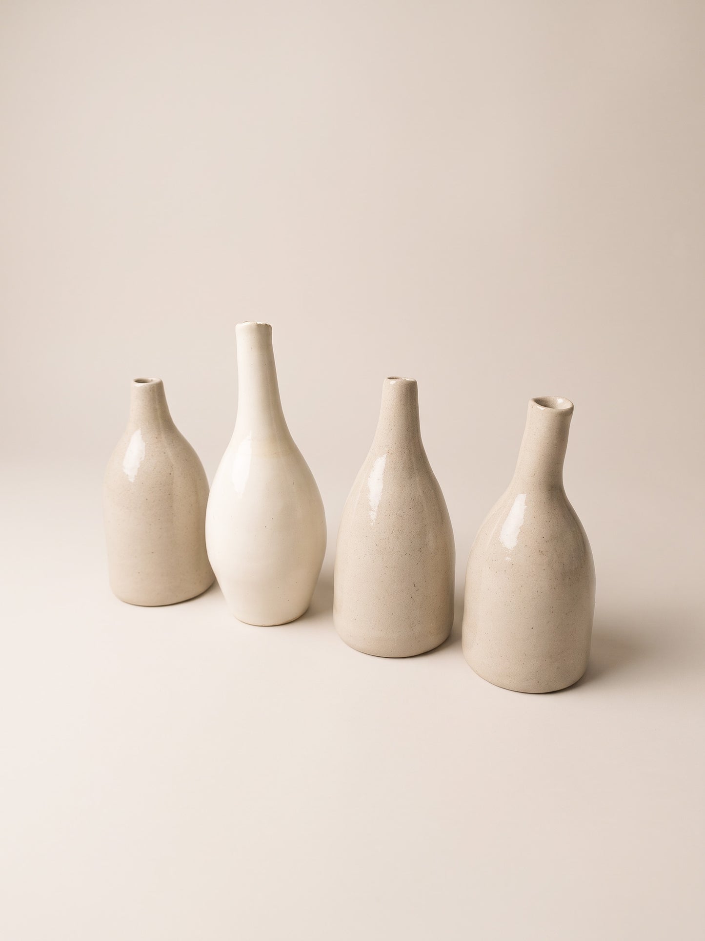Four minimalist ceramic vases in warm off-white and cream tones, arranged in a row against a solid, light beige background.