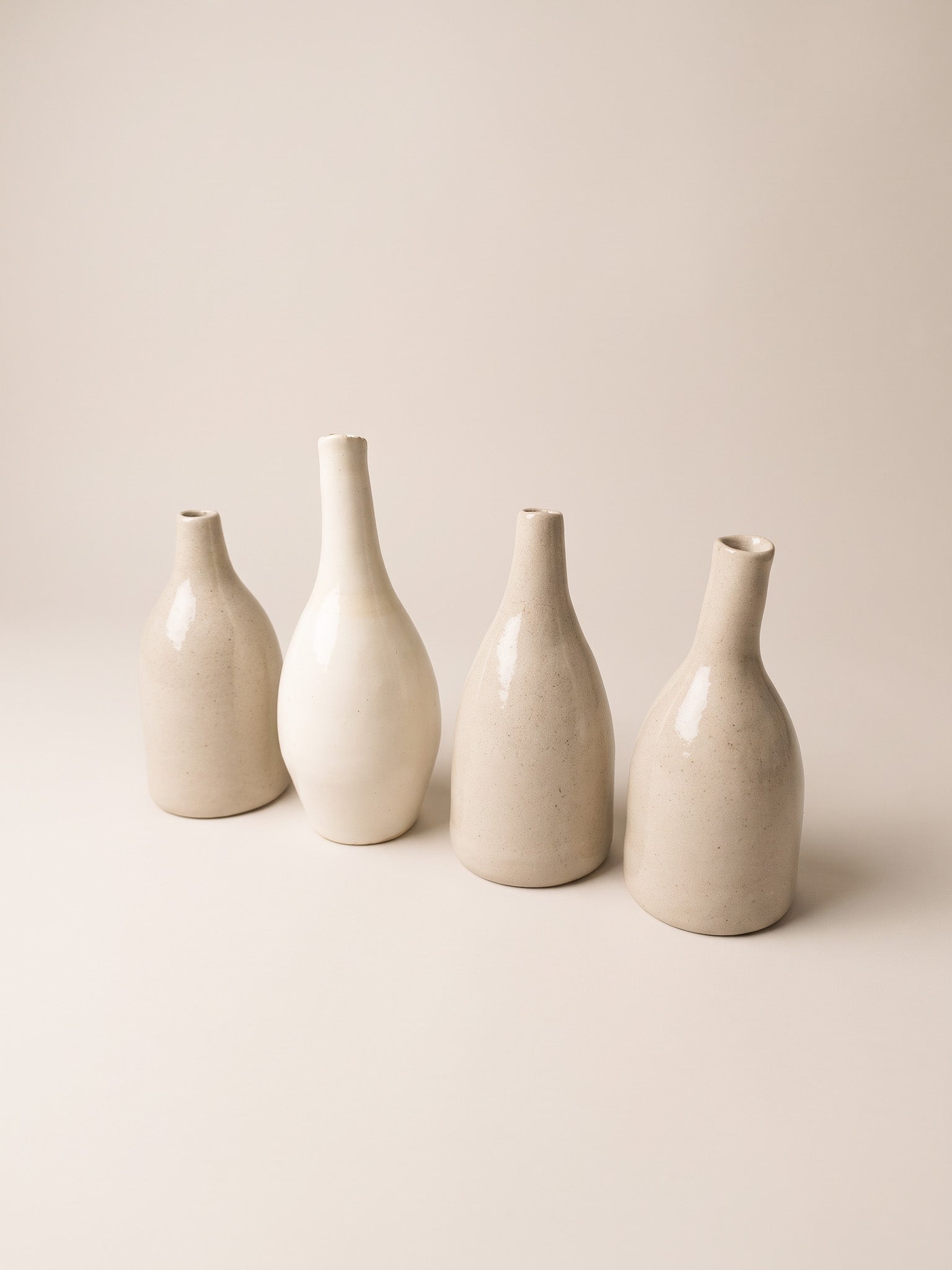 Four minimalist ceramic vases in warm off-white and cream tones, arranged in a row against a solid, light beige background.