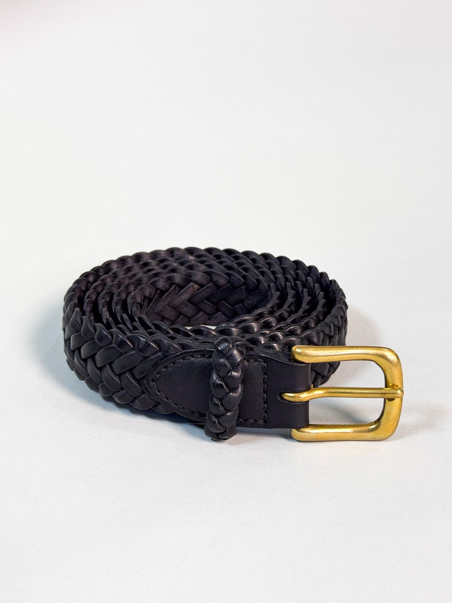 A black braided leather belt with a polished gold-tone buckle, coiled on a light gray background.