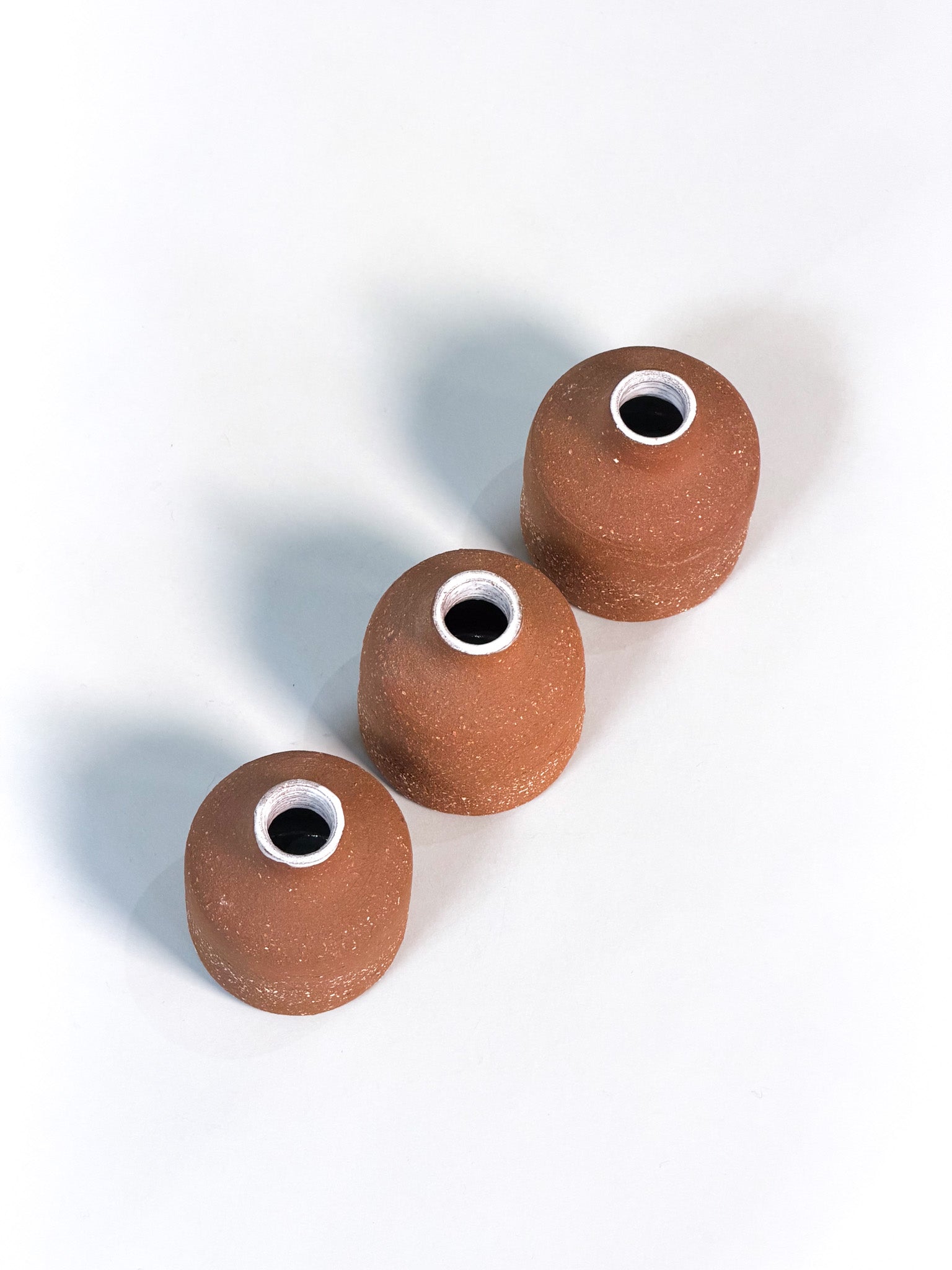 Three small terracotta-colored ceramic vases with a matte, speckled finish and a white inner rim around the opening, arranged diagonally on a white background.