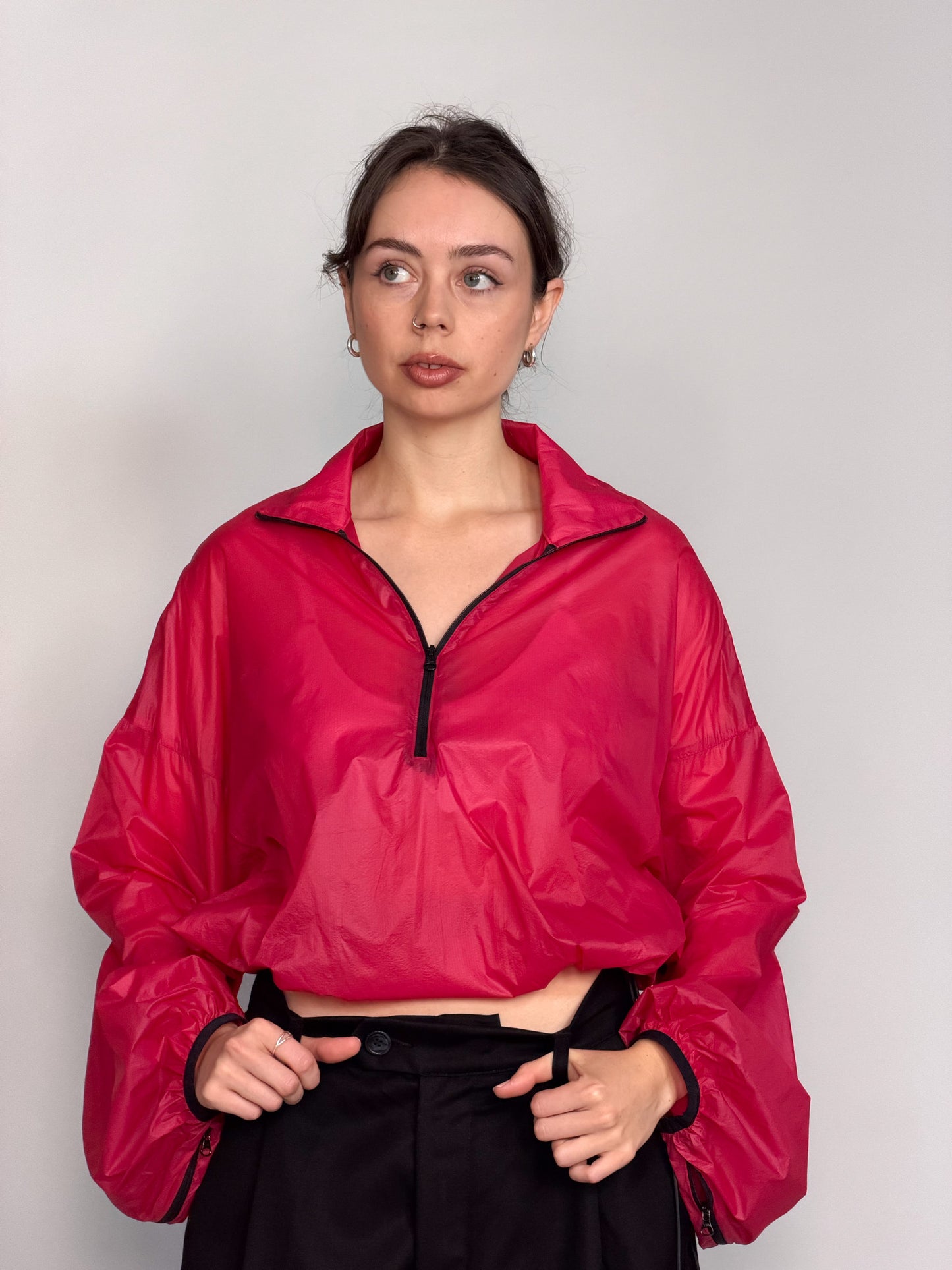 A woman modeling a bright pink semi-sheer zip-up windbreaker jacket with voluminous balloon sleeves, paired with black wide-leg trousers, against a light gray studio background.