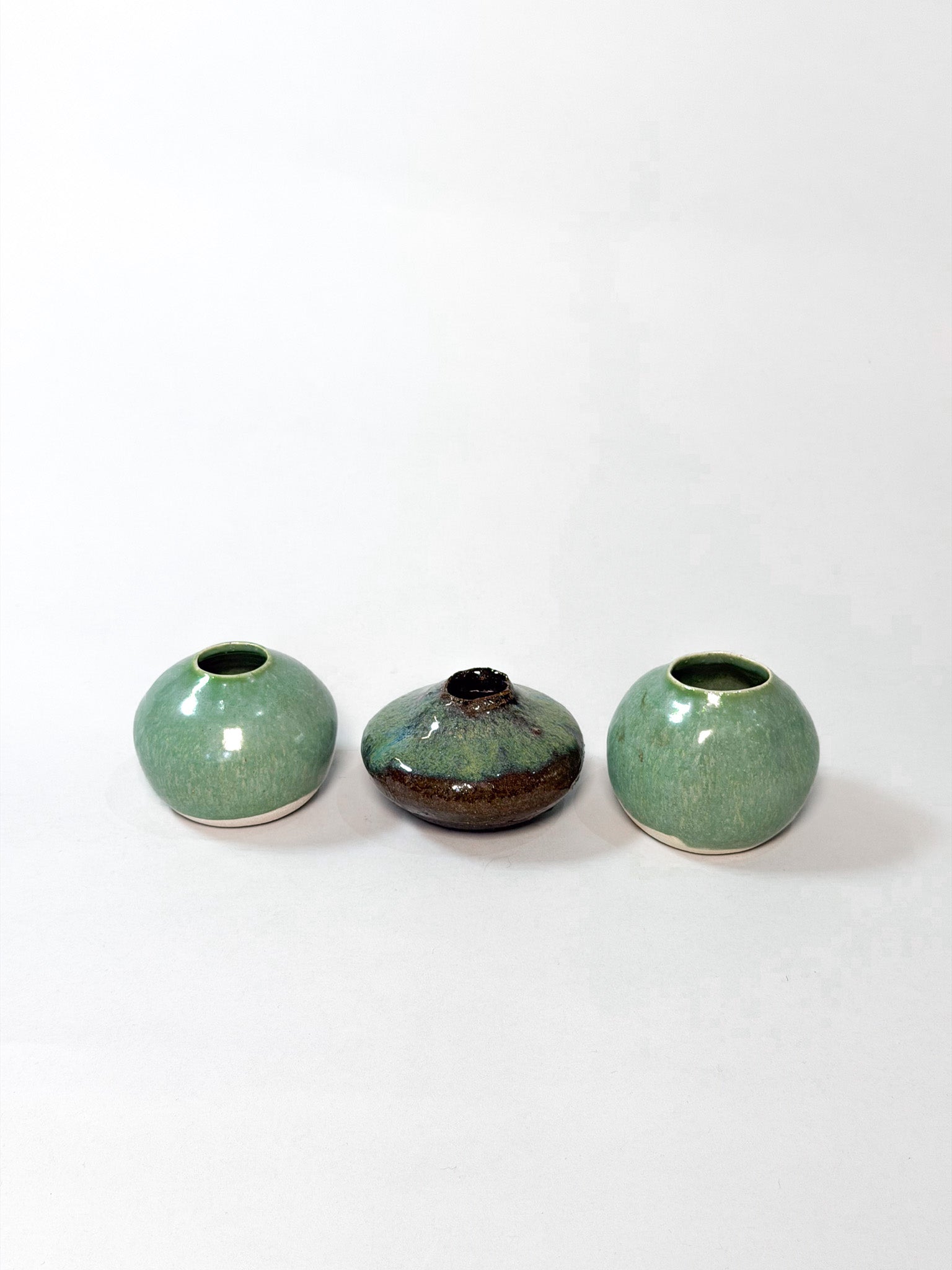Three small ceramic vases arranged in a row on a plain white background. The two outer vases are rounded and glazed in a light green glaze, while the center vase is darker with a brown and black mottled finish.