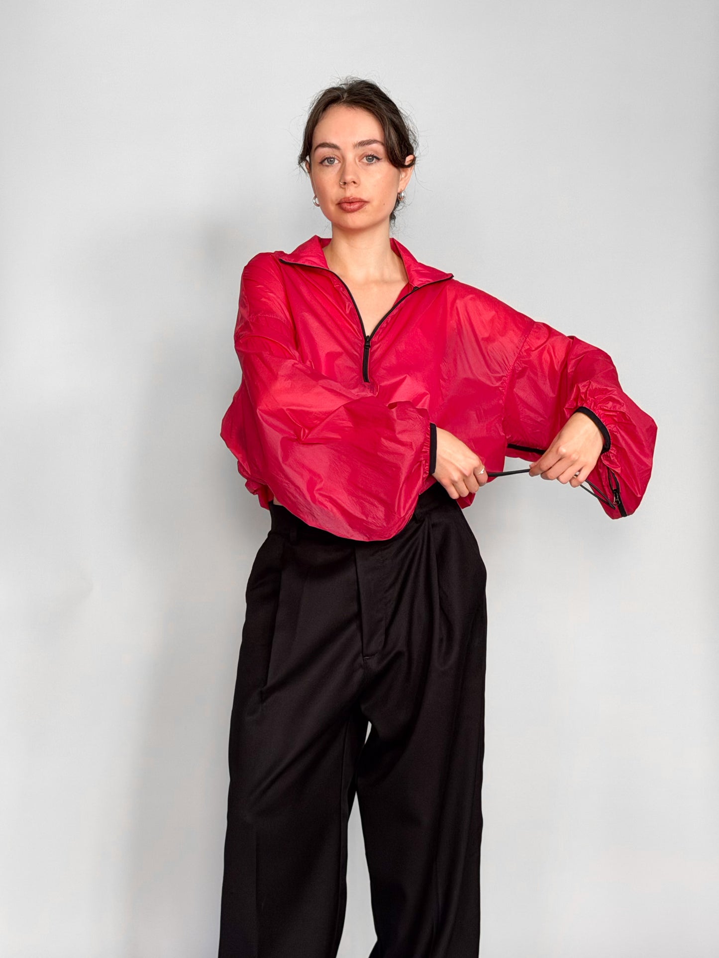 A woman modeling a bright pink semi-sheer zip-up windbreaker jacket with voluminous balloon sleeves, paired with black wide-leg trousers, against a light gray studio background.