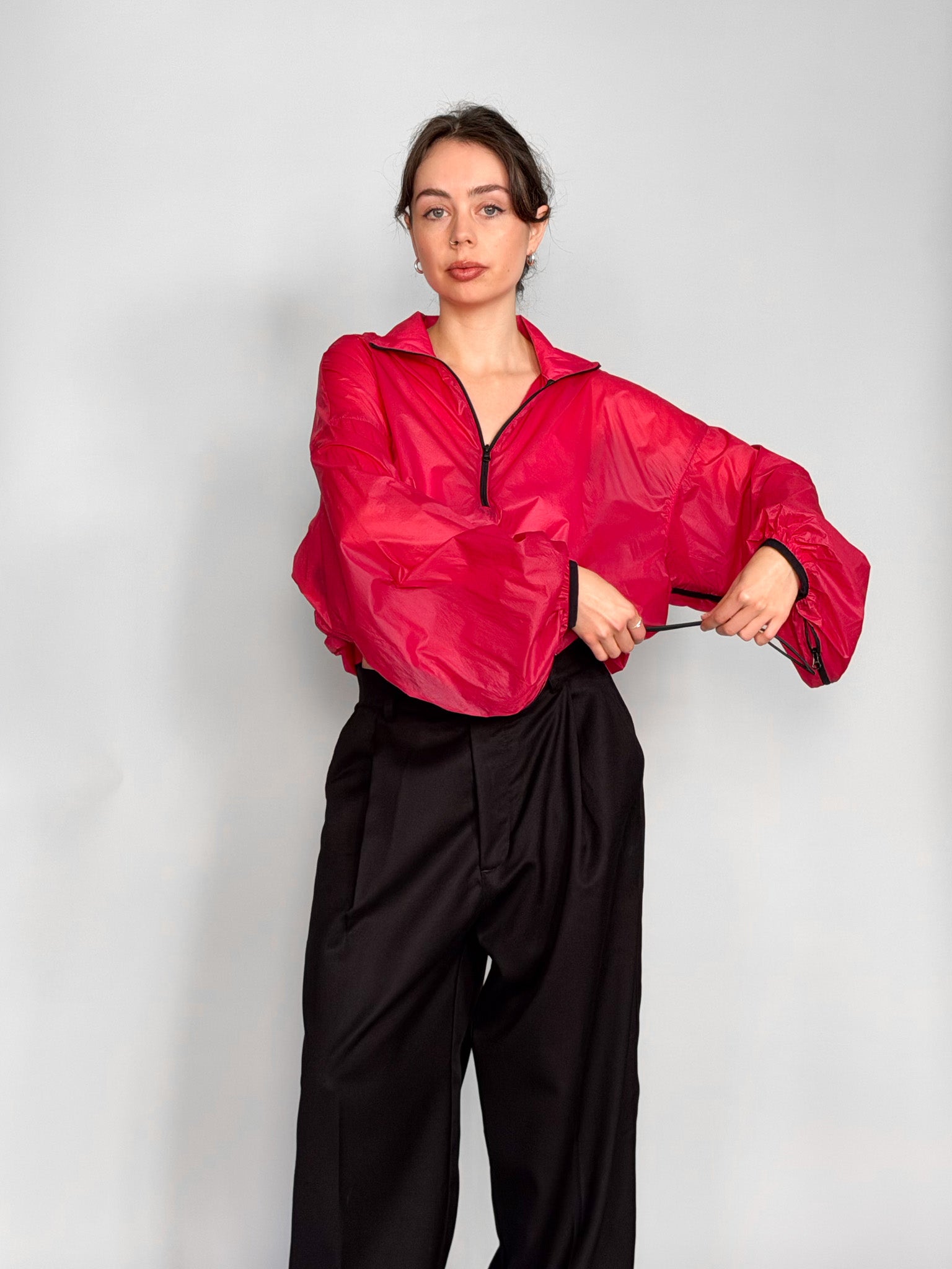 A woman modeling a bright pink semi-sheer zip-up windbreaker jacket with voluminous balloon sleeves, paired with black wide-leg trousers, against a light gray studio background.