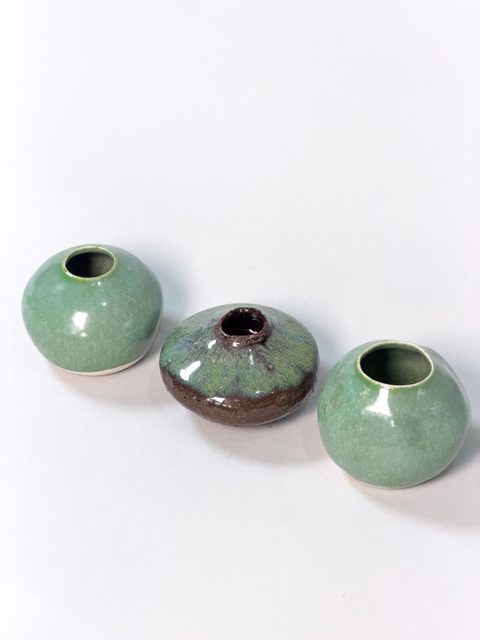 Three small handmade ceramic vases with a glossy glaze, arranged on a plain white background. Two vases are light green, and the center vase has a mottled green and dark brown glaze.