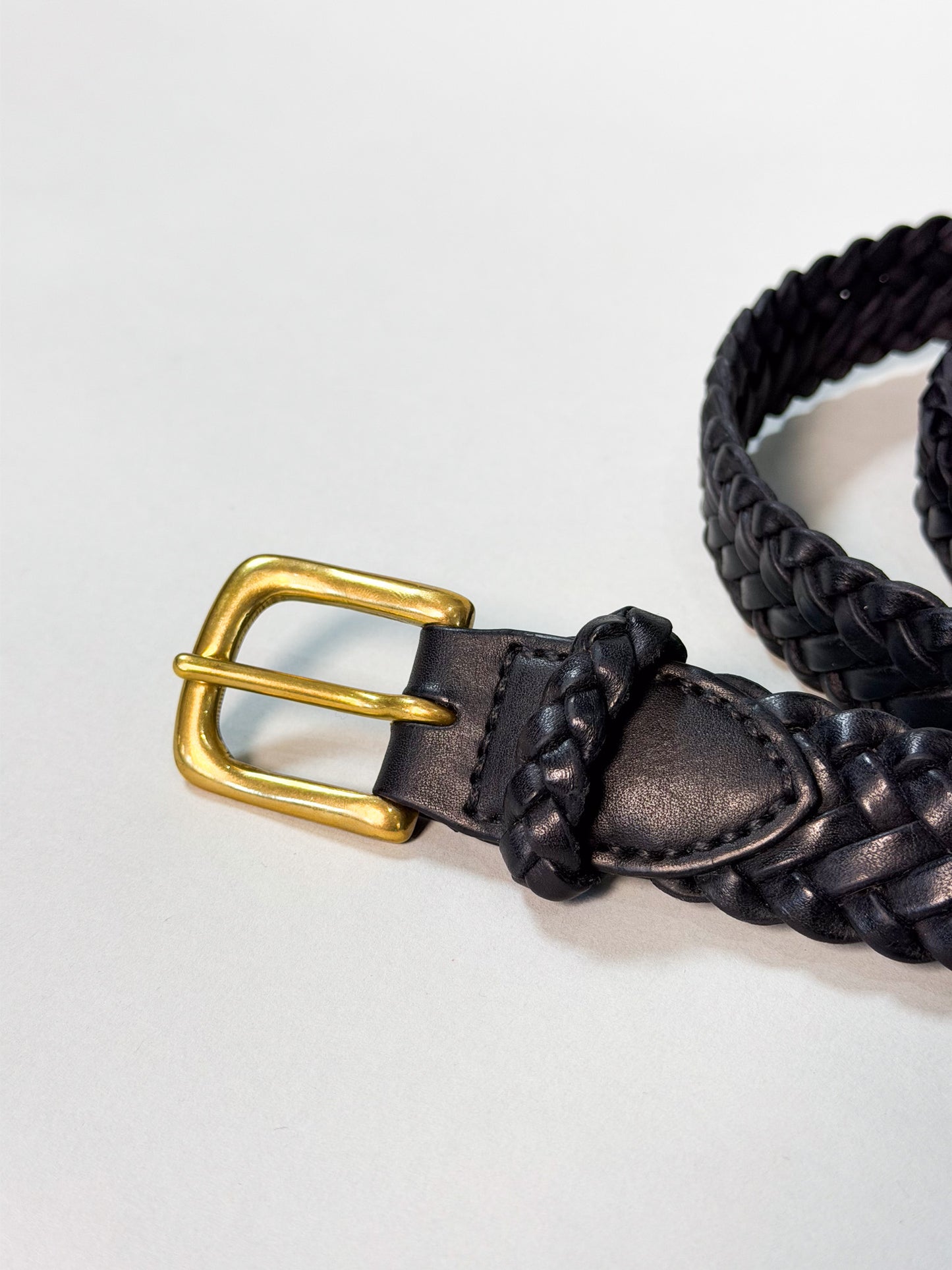 A close-up of a black braided leather belt with a polished gold-tone metal buckle, resting on a light gray background.