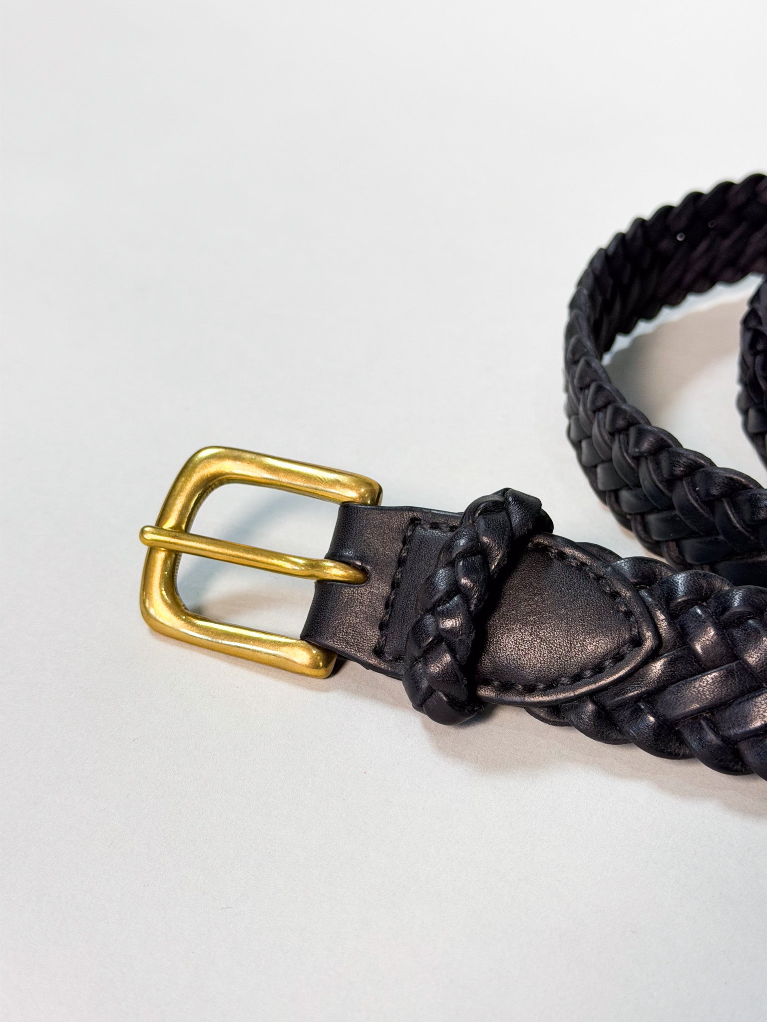 A close-up of a black braided leather belt with a polished gold-tone metal buckle, resting on a light gray background.