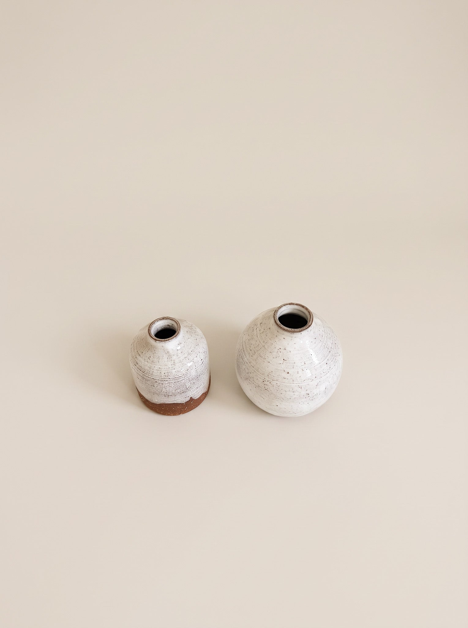 Two small ceramic vases with a speckled white and gray glaze and a natural brown clay base, displayed on a solid beige background.