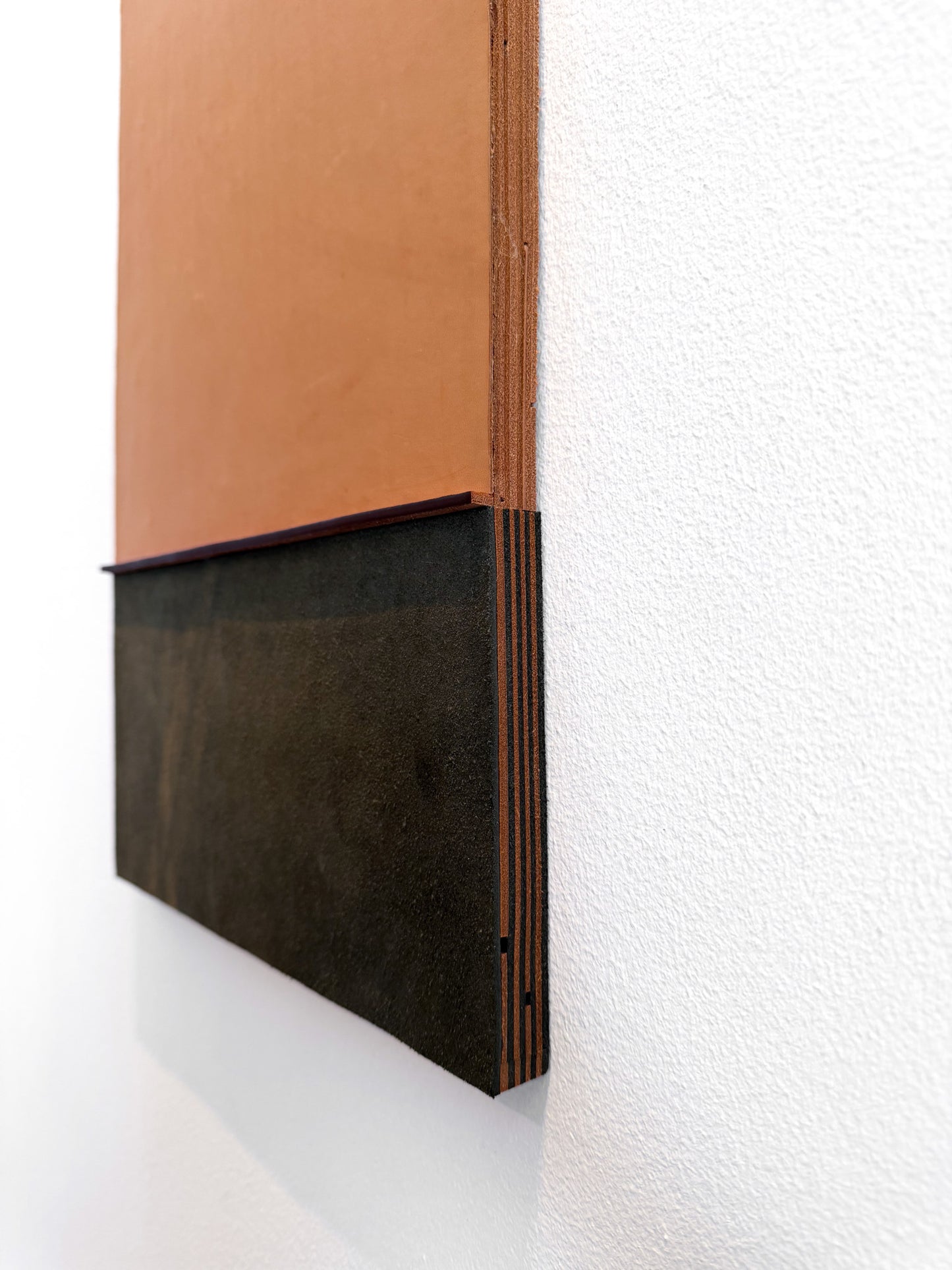 A close-up of a modern wall-mounted art piece featuring a terracotta orange upper section and a dark charcoal-brown textured lower section, mounted on a white textured wall.