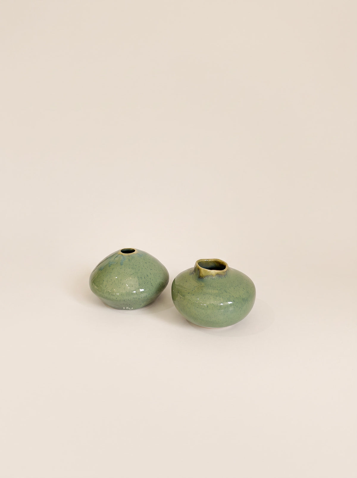 Two small rounded ceramic vases with a glossy green glaze and subtle gold-toned highlights, shown on a plain warm beige background.