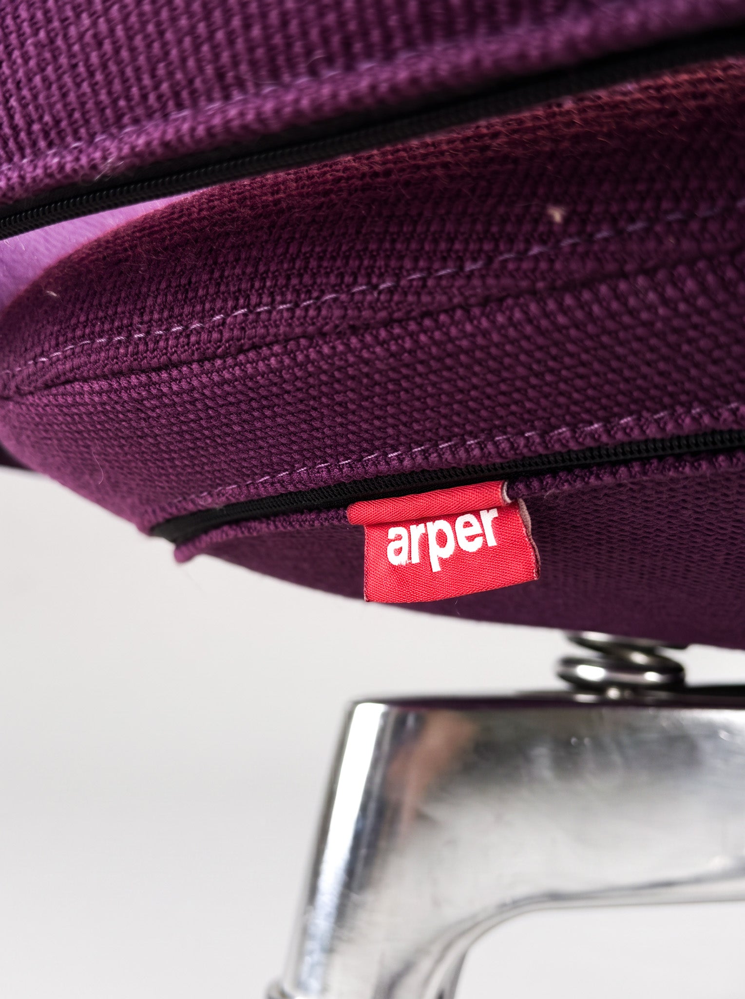 Close-up of a purple woven fabric product with a black zipper and a red Arper brand tag, shown against a white background with part of a metallic silver object visible below.