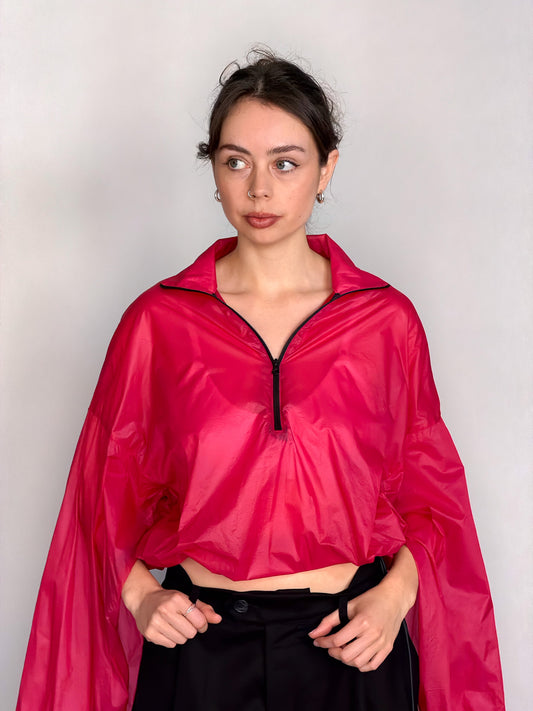 A woman modeling a bright pink semi-sheer zip-up windbreaker jacket with voluminous balloon sleeves, paired with black wide-leg trousers, against a light gray studio background.